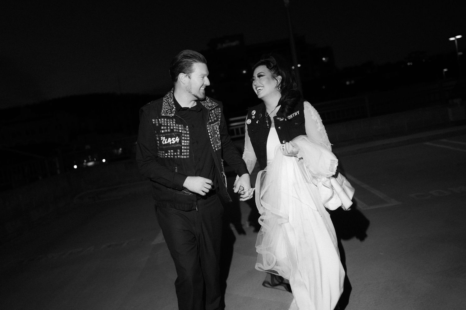 A couple in contrasting wedding and punk attire walk together in a dark parking area.