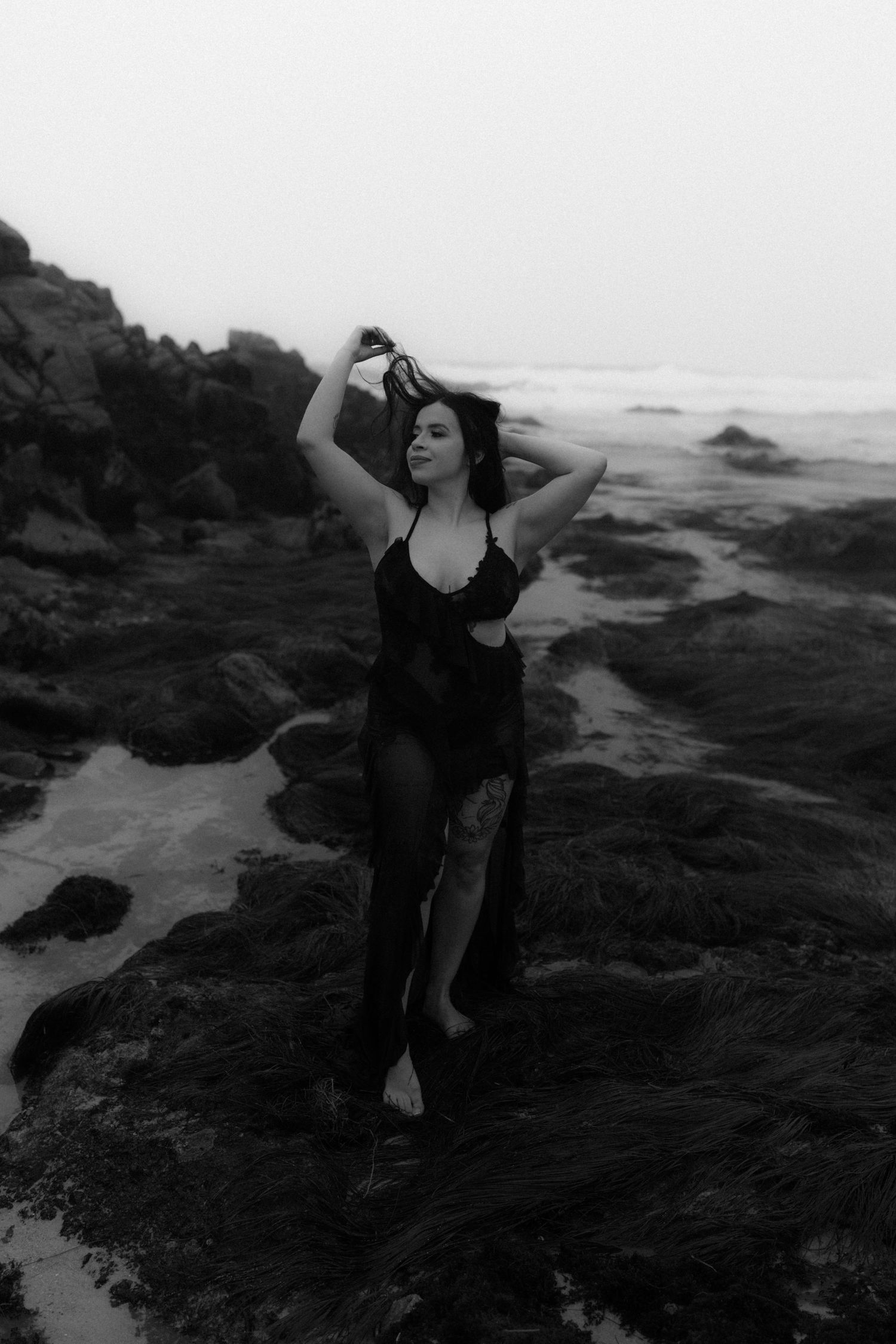 Monochrome coastal portrait with figure in dark dress on rocky shore.