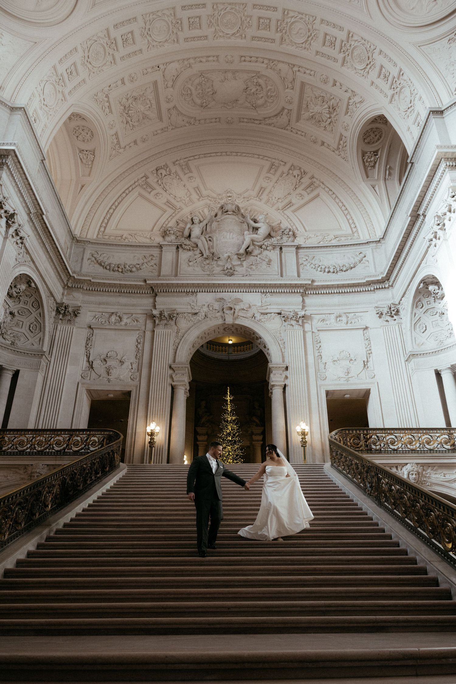 A couple walks up an ornate grand staircase in a baroque palace with white walls and decorative moldings.