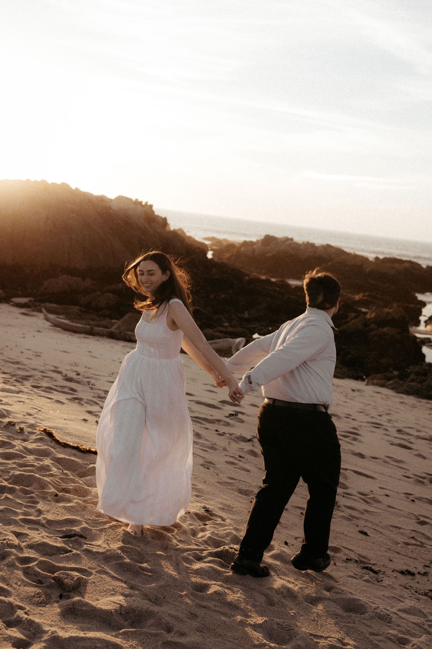 A couple dances together on a sandy beach at sunset, with the woman wearing a flowing white dress.
