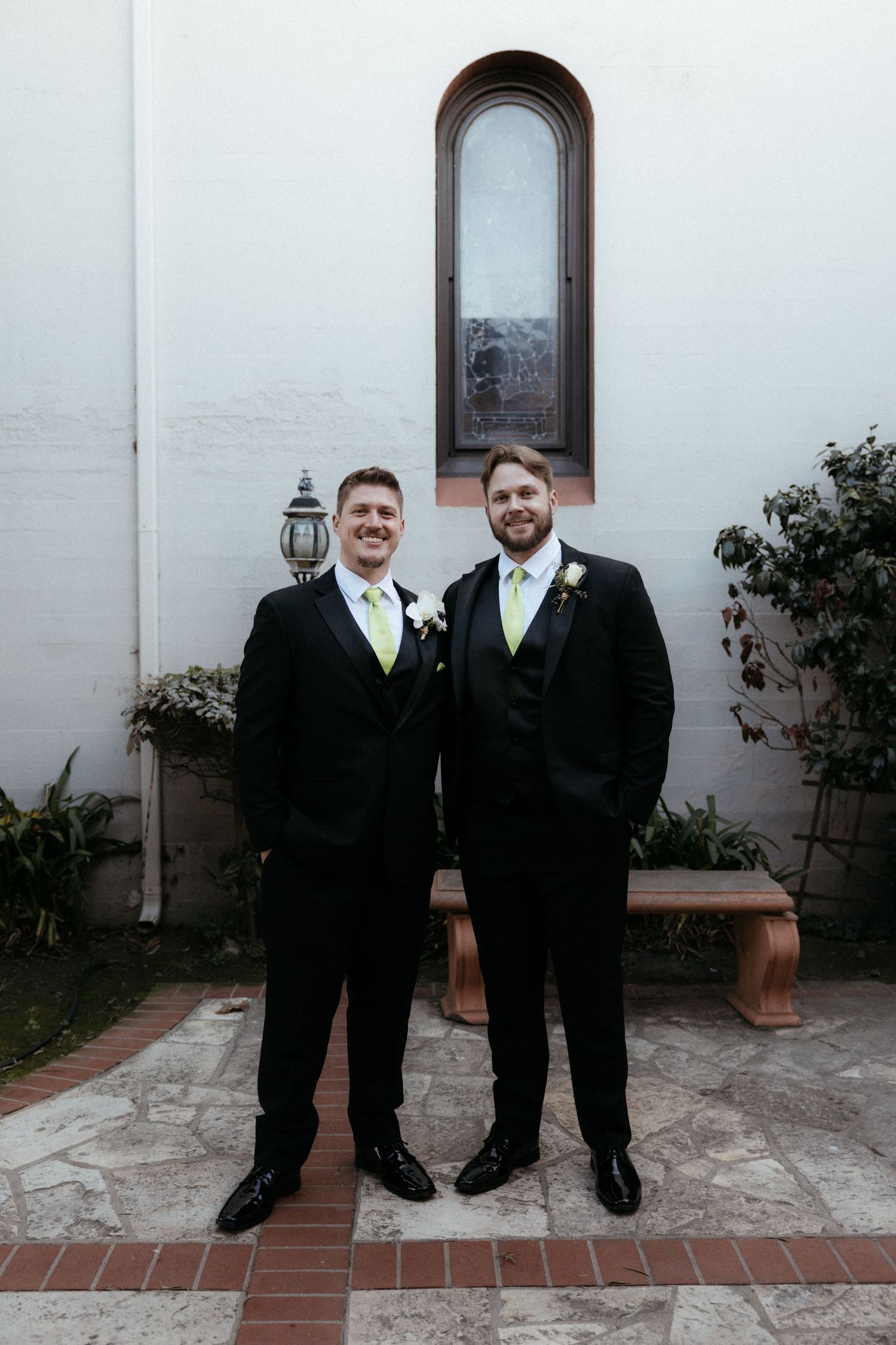 Two groomsmen in black suits with green ties stand together on a brick patio outside a white building.