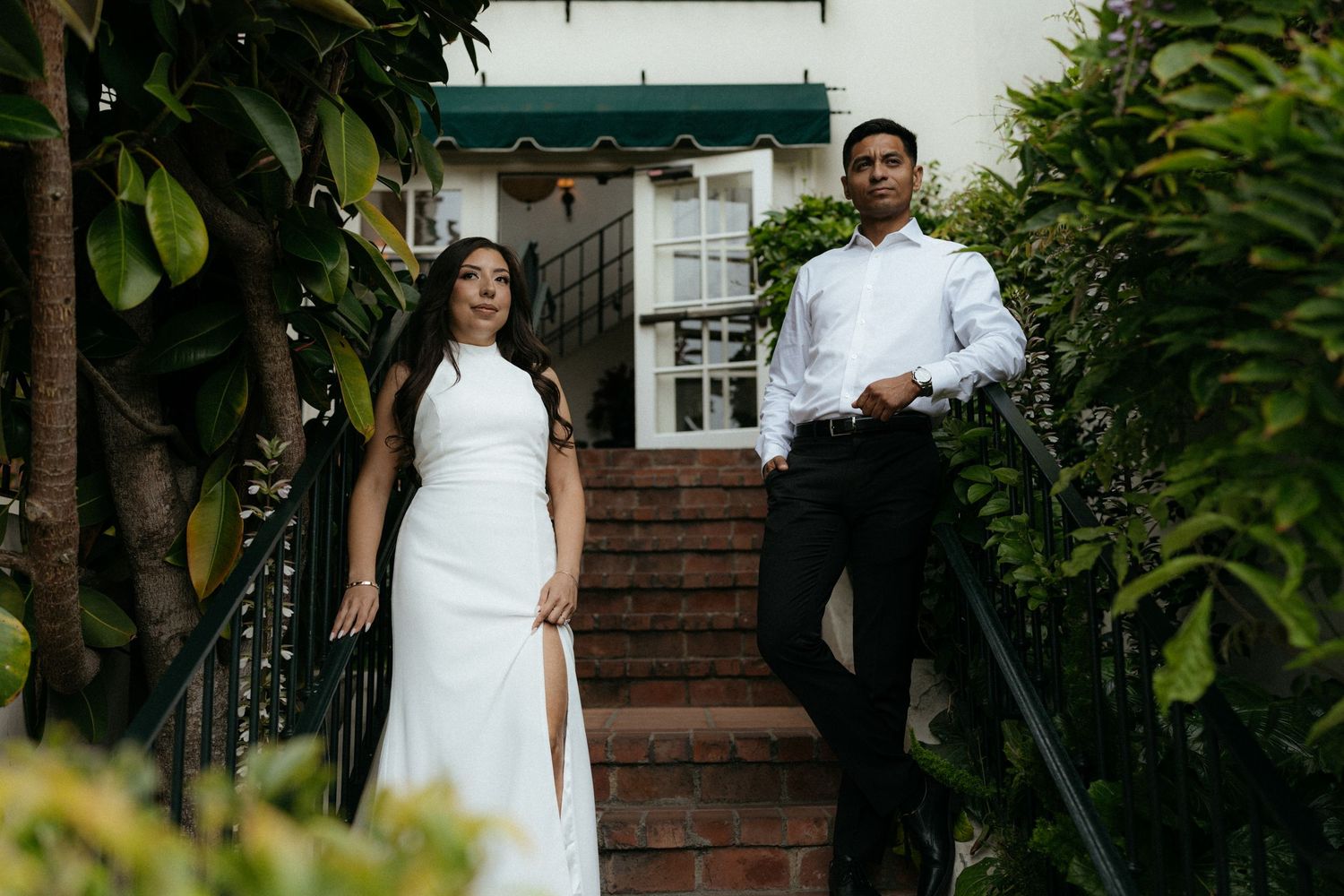 Couple in formal attire posing on brick stairs surrounded by lush greenery, woman in white gown, man in black trousers.