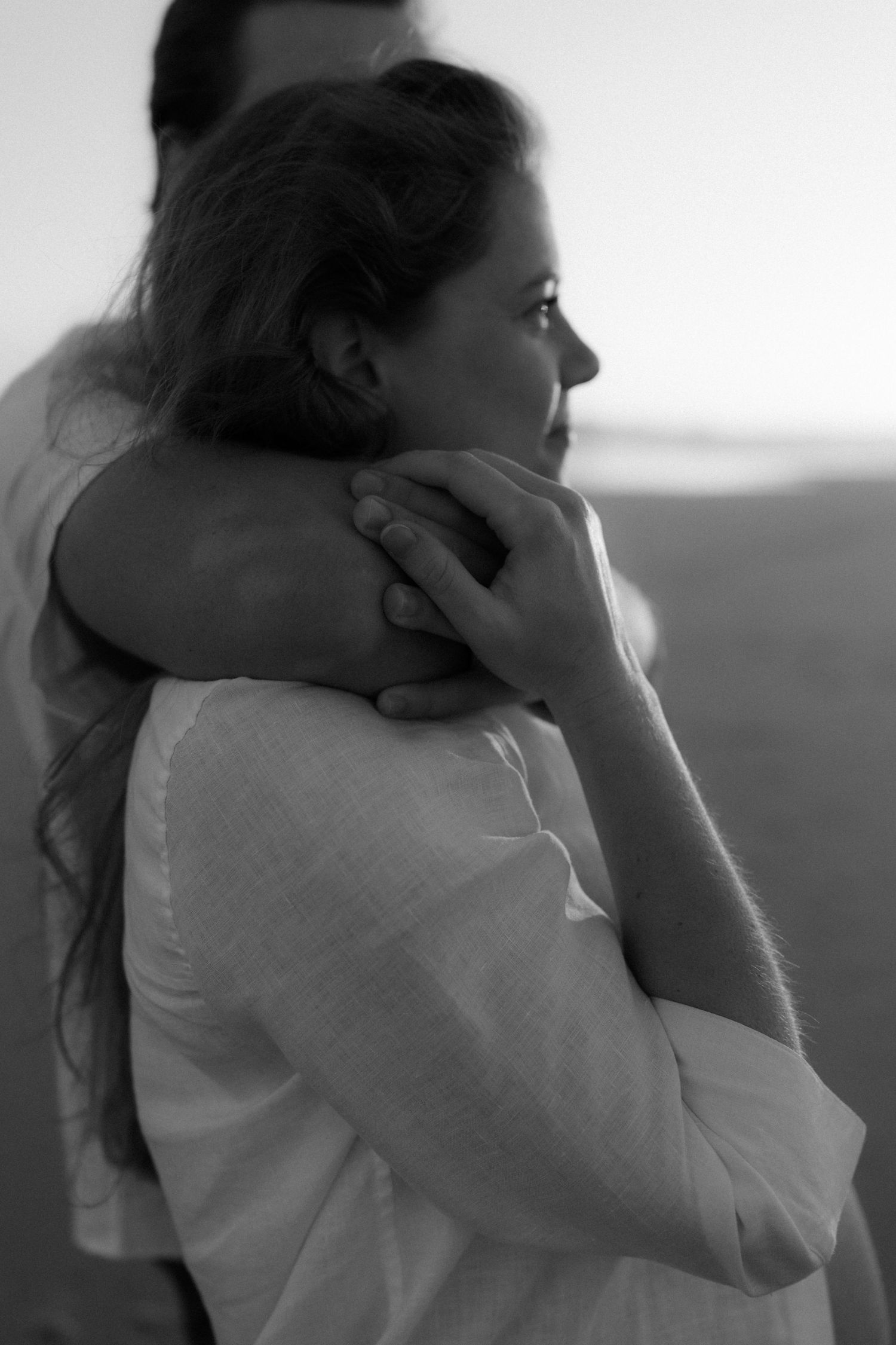 Black and white photo of a couple embracing outdoors, woman in white linen shirt gazing into distance.