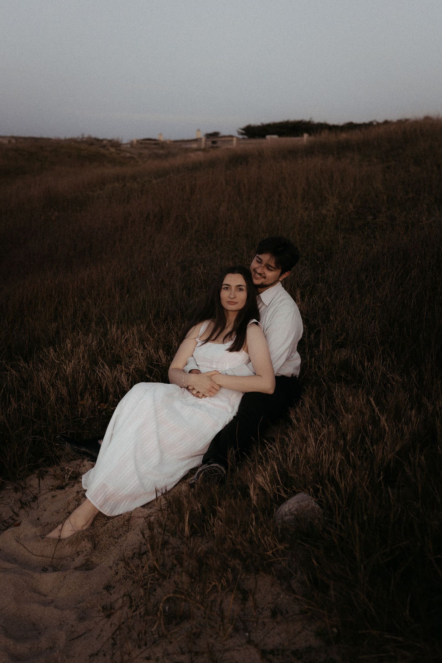 Romantic couple sitting together in coastal grass dunes at dusk, woman in white dress.