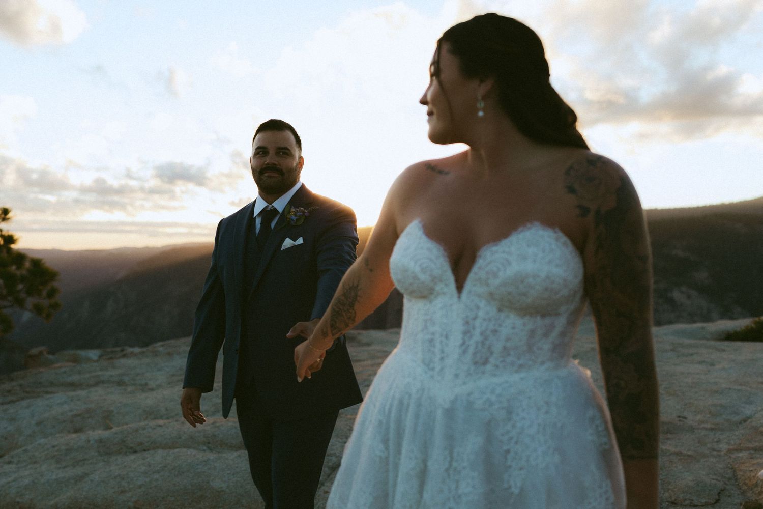 Bride and groom hold hands at sunset on a mountain overlook, bride in lace gown with tattoos.