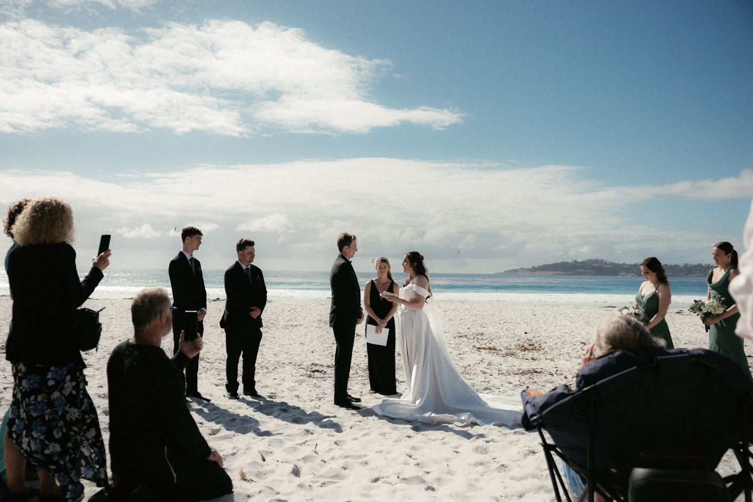 A couple exchanges wedding vows on a white sandy beach with guests watching under a blue sky.