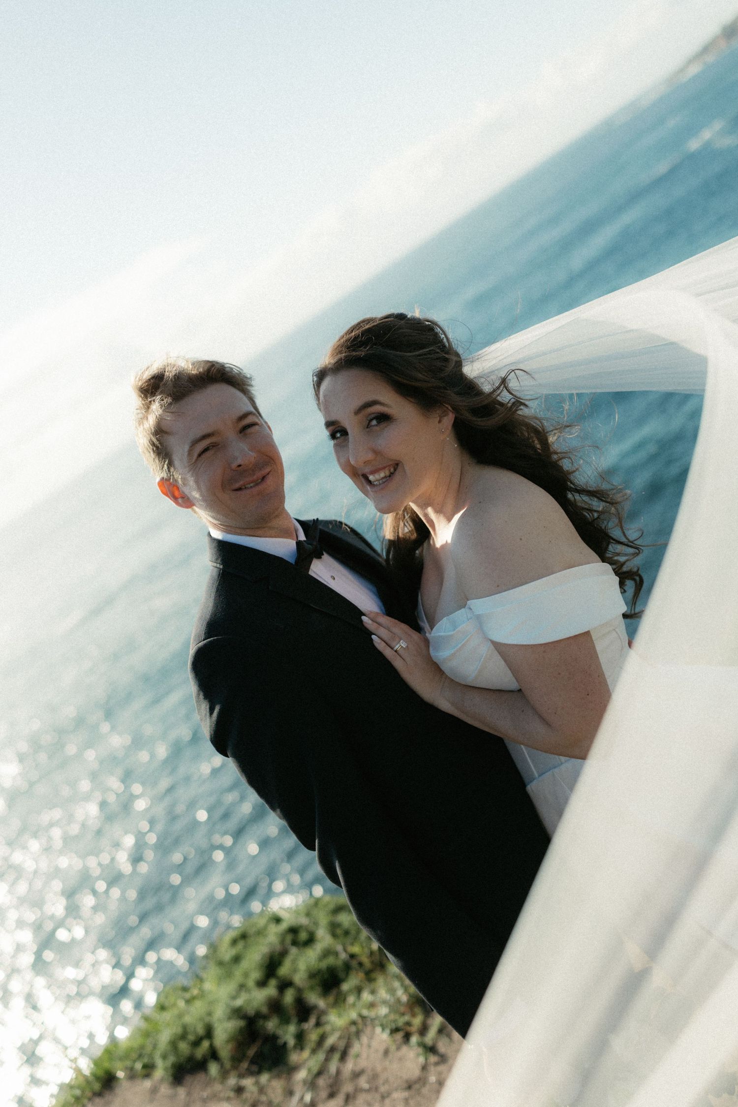 Bride and groom smiling together by the ocean, bride in off-shoulder white gown with veil flowing in the breeze.