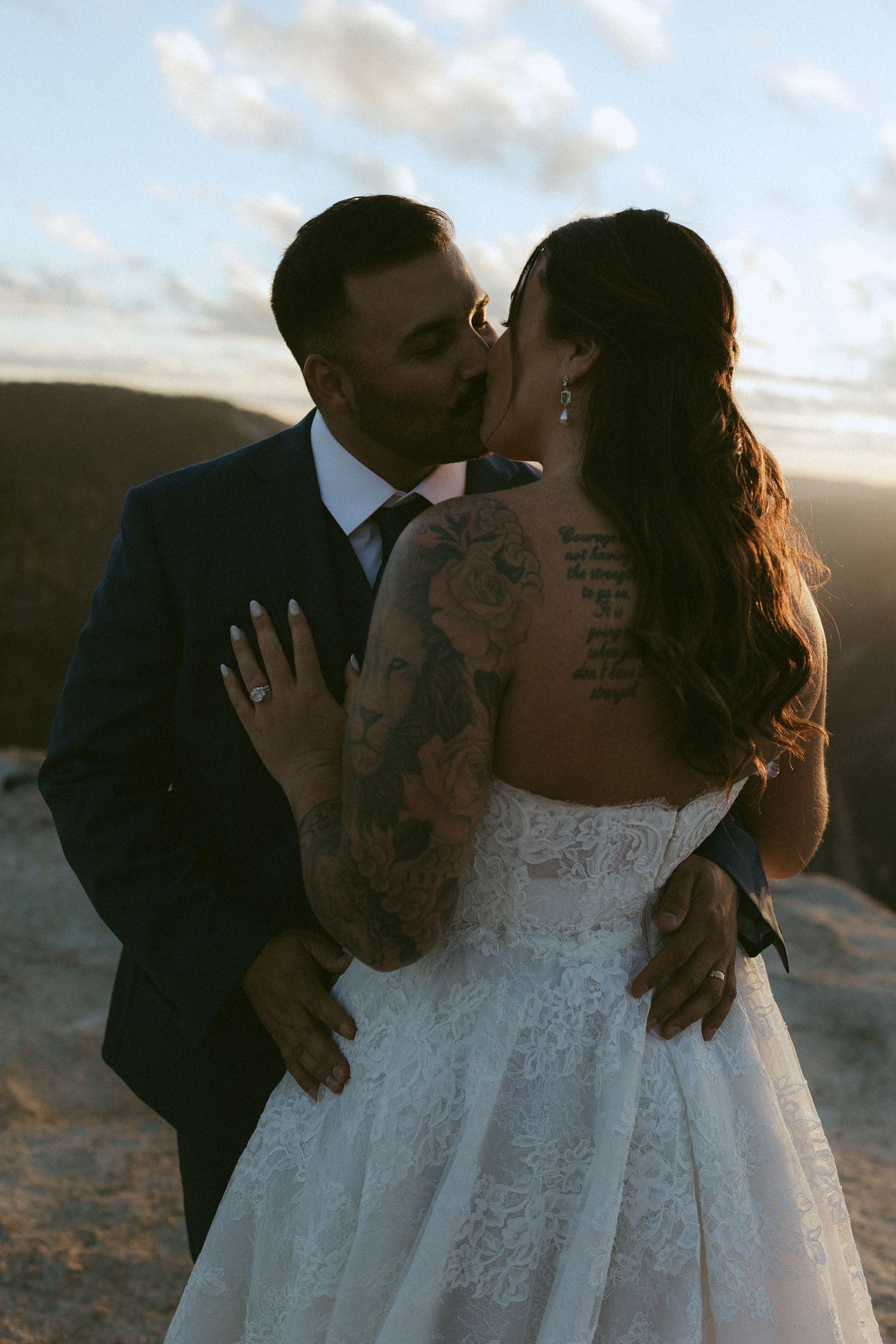 Bride with floral arm tattoos kisses groom at sunset on mountain overlook.