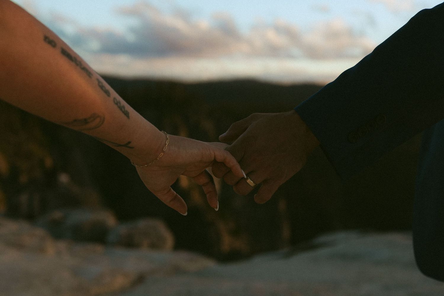 Two hands gently touching, one with tattoos and a ring, against a scenic sunset backdrop outdoors.