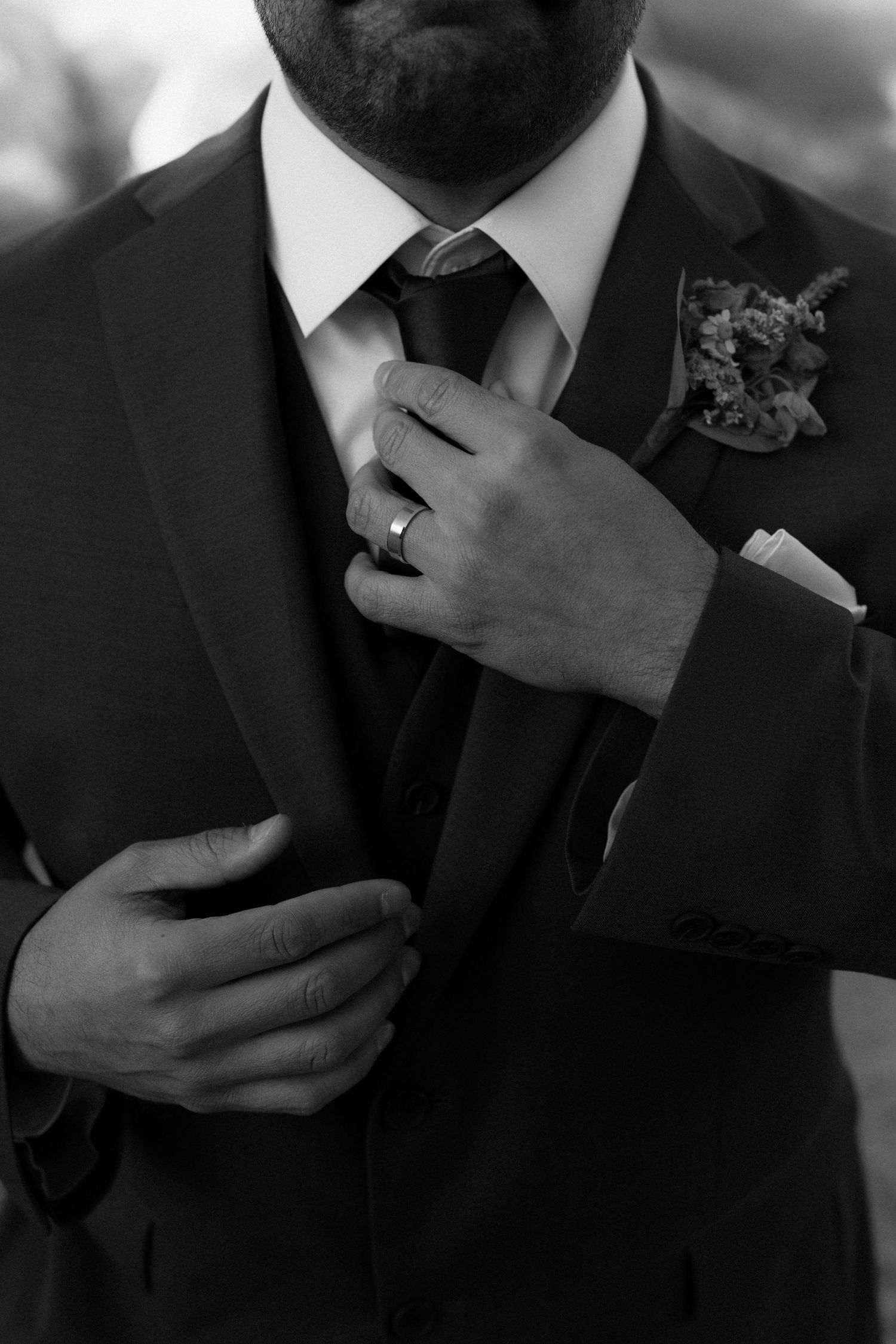 Groom in dark suit adjusting tie with boutonniere and pocket square in black and white.