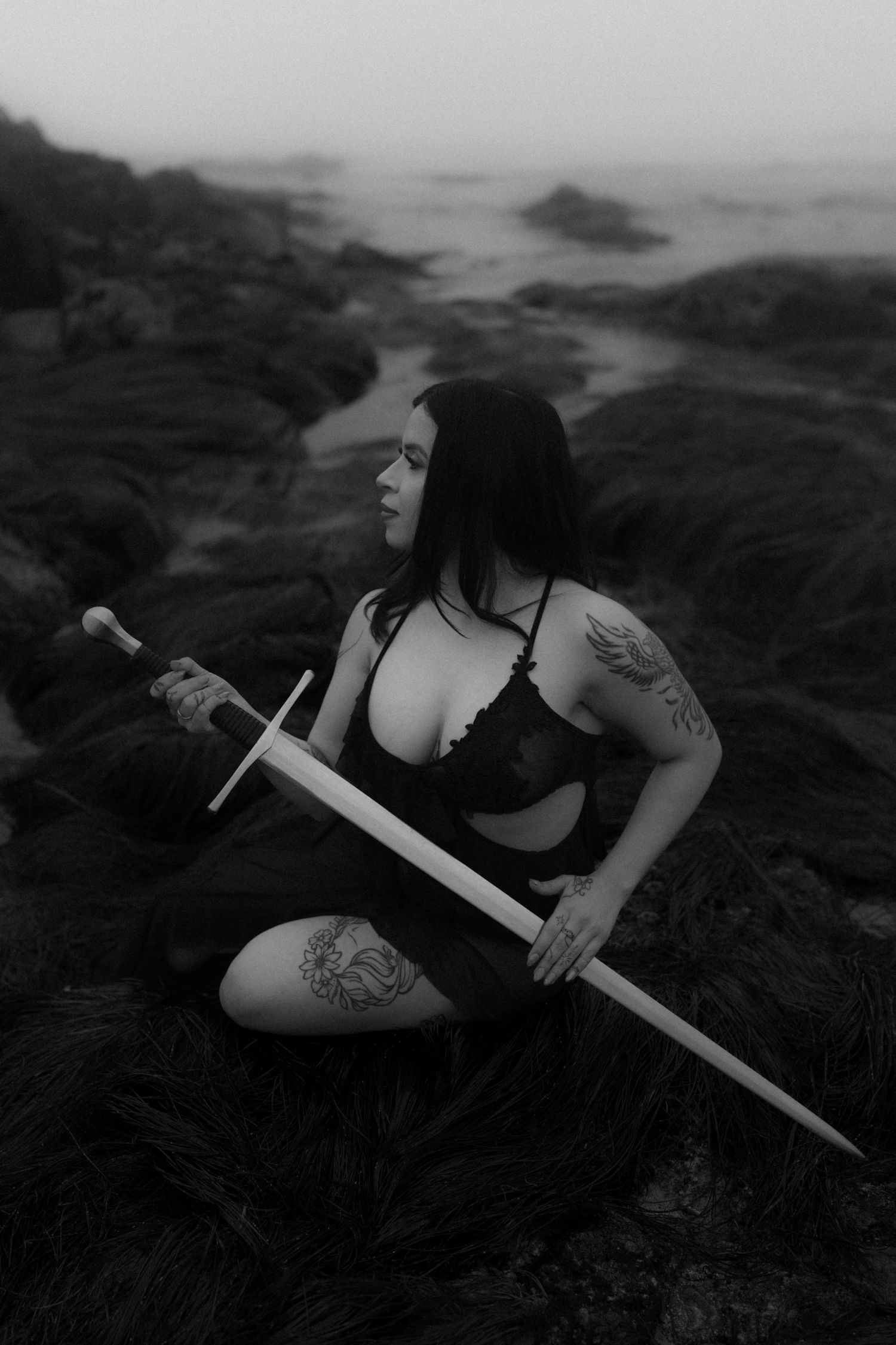 Monochromatic image of someone posing with sword on dark rocky shore.