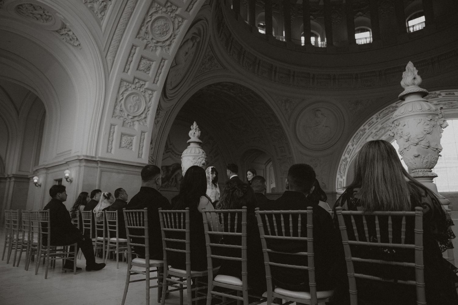 A wedding ceremony in a grand ornate hall with white chairs and architectural details.