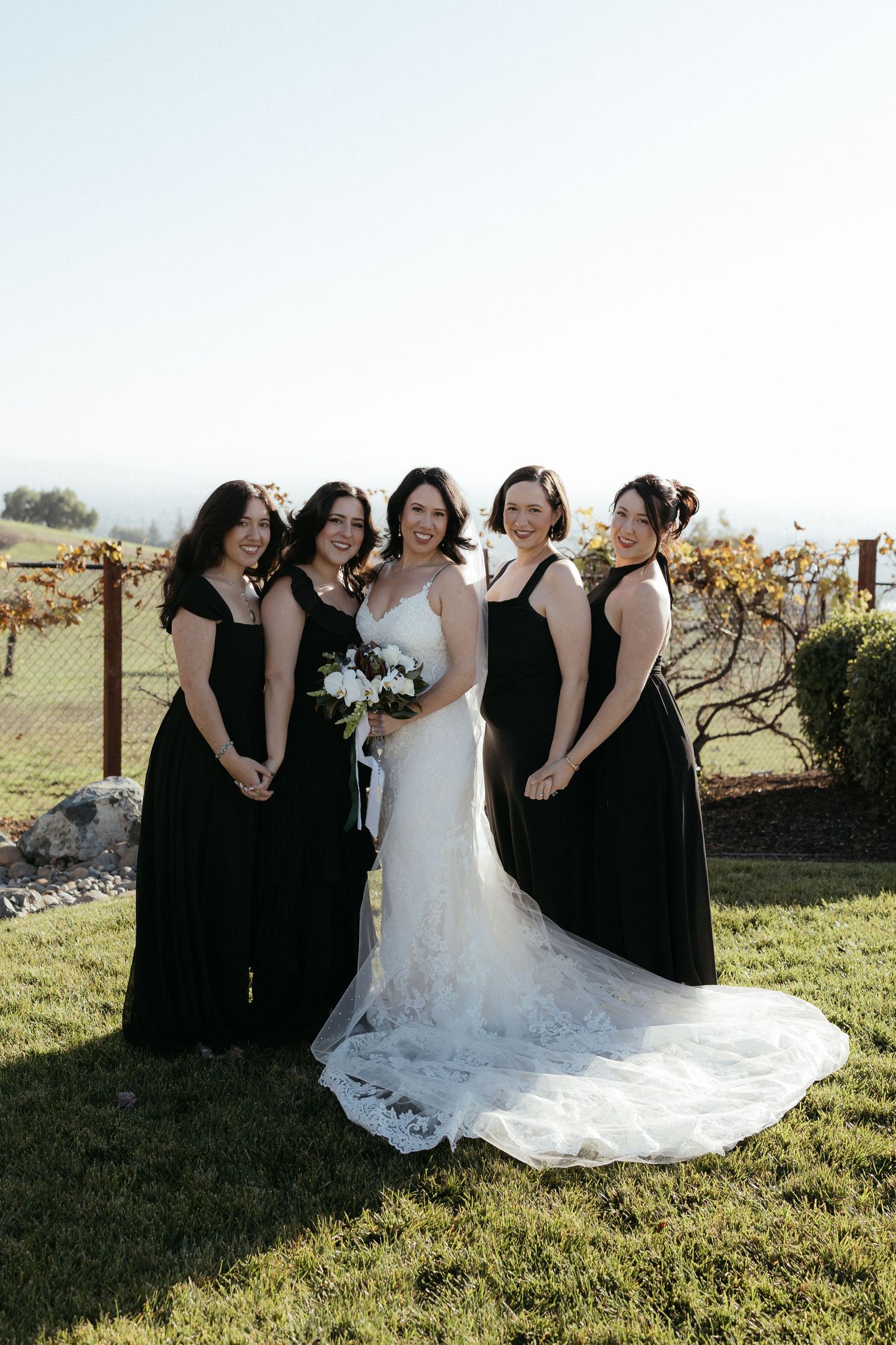 A bride in a white lace gown poses with bridesmaids wearing black dresses in a sunny outdoor setting.