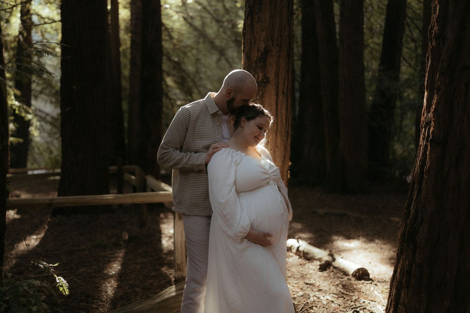 Dancing couple in white and beige outfits embrace among towering redwood trees in dappled forest light.