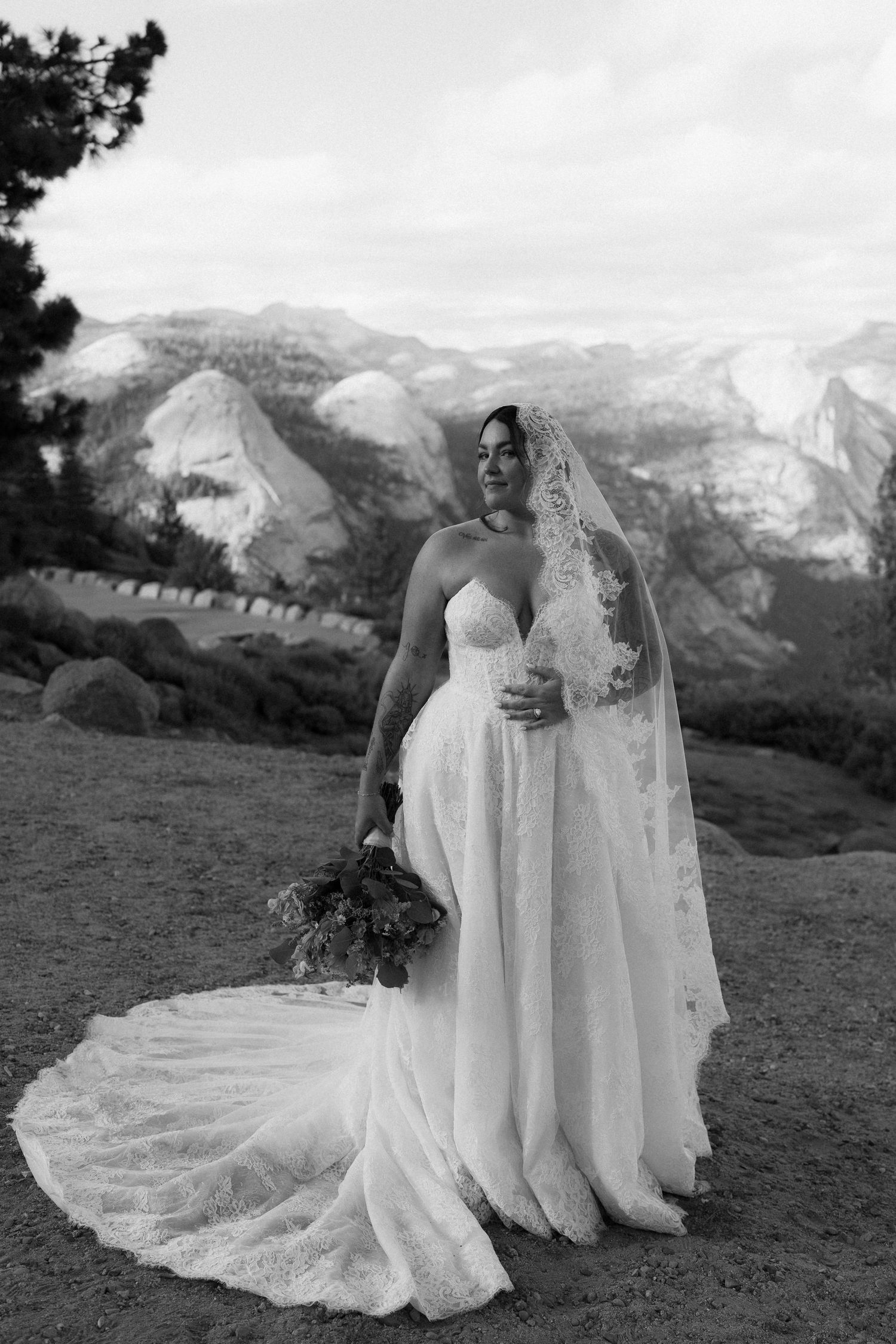 Bride in lace wedding gown and veil holding bouquet with Yosemite mountains backdrop.