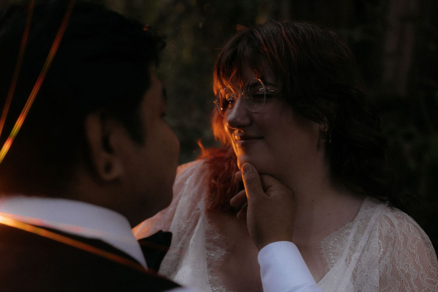 Groom gently touches bride's face during romantic outdoor wedding ceremony in soft warm evening light.