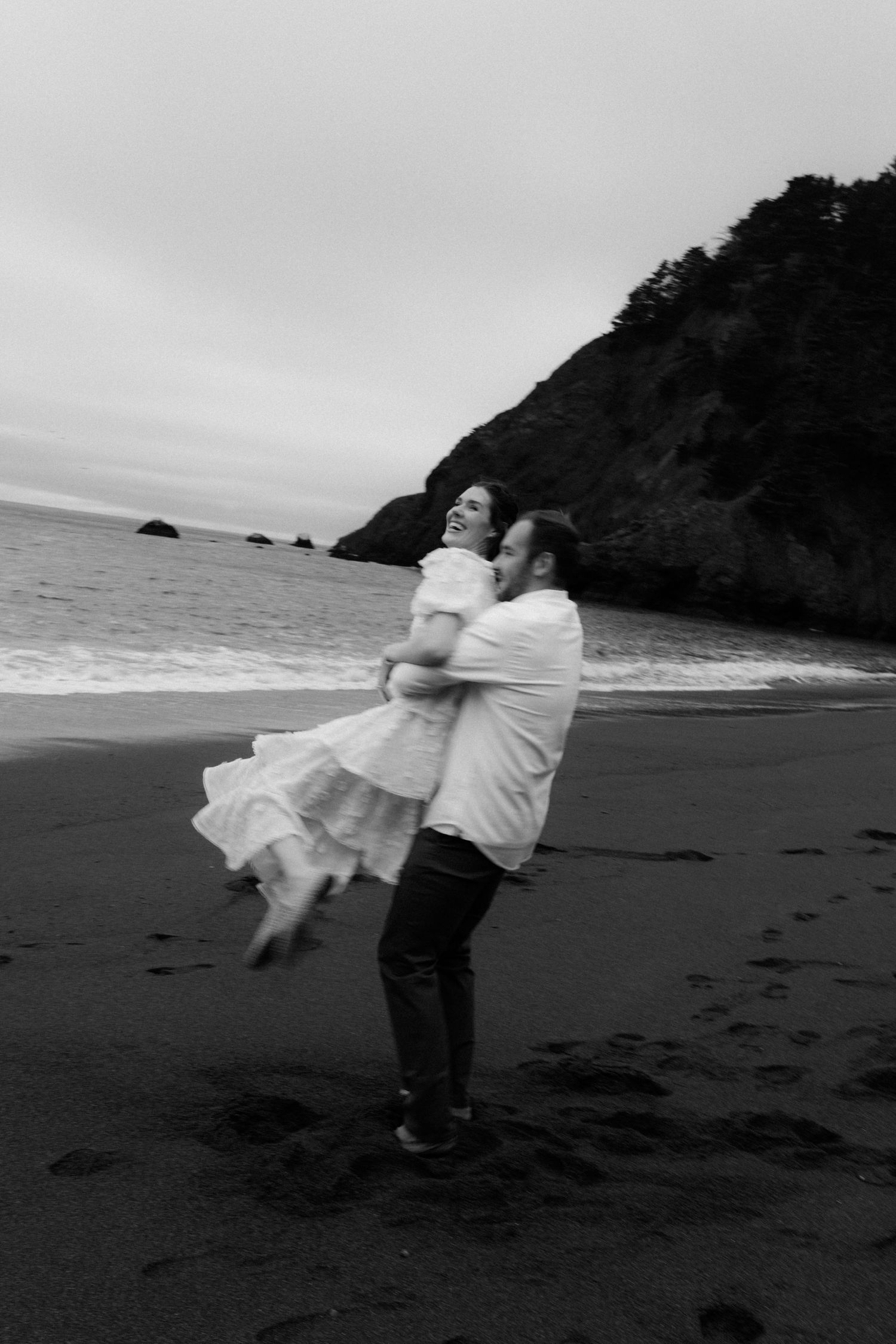 Black and white photo of a couple embracing and laughing on a beach, man lifting woman in white dress near ocean.