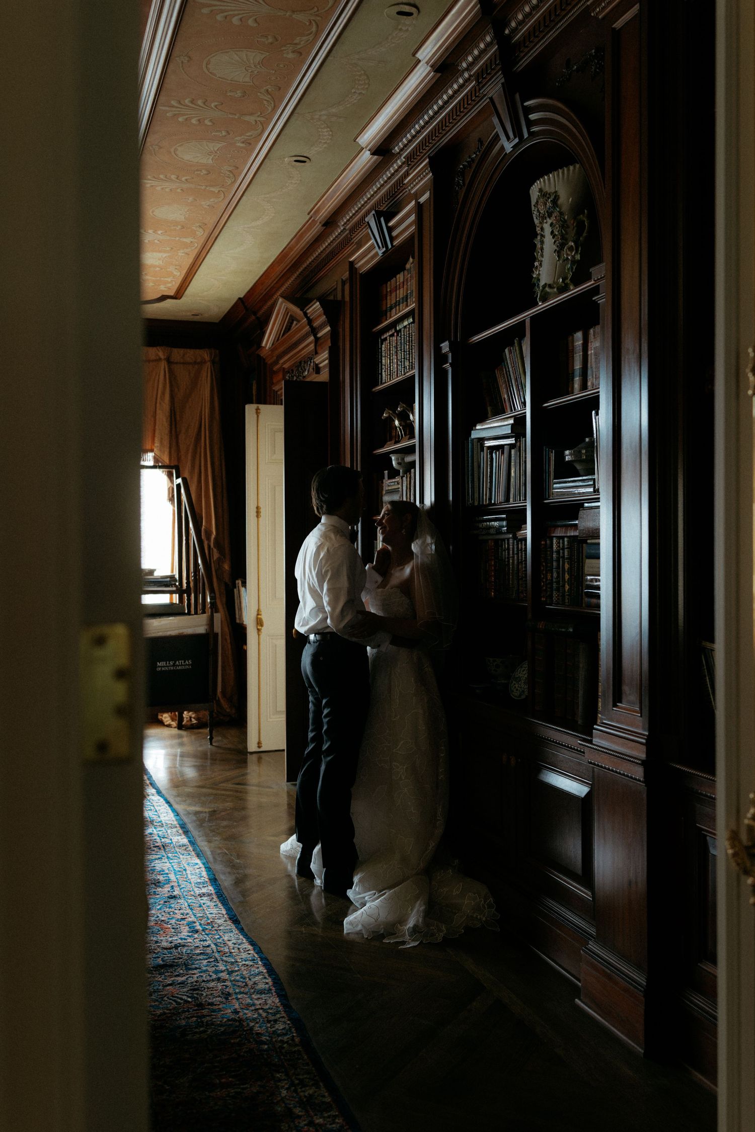 A bride and groom share an intimate moment in a dark, elegant wood-paneled library with floor-to-ceiling bookshelves.