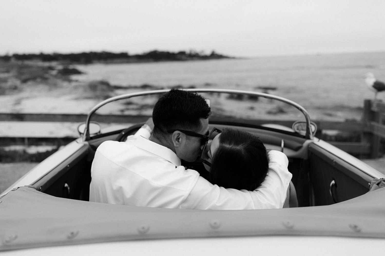 Black and white photo of a couple kissing in a vintage convertible car by the ocean.