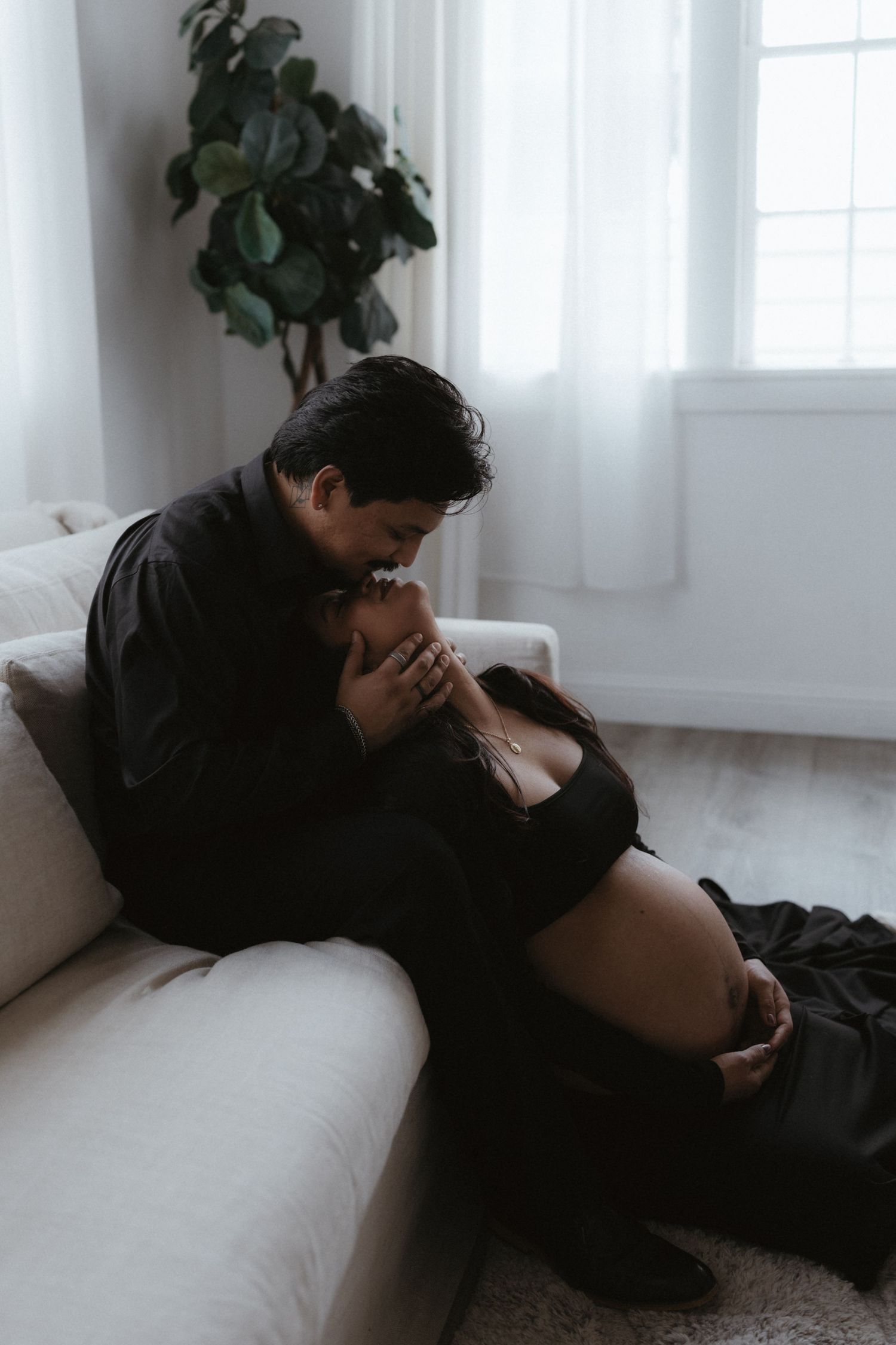 Couple in black outfits sharing an intimate moment on a white sofa, with a pregnant belly visible in a softly lit room.