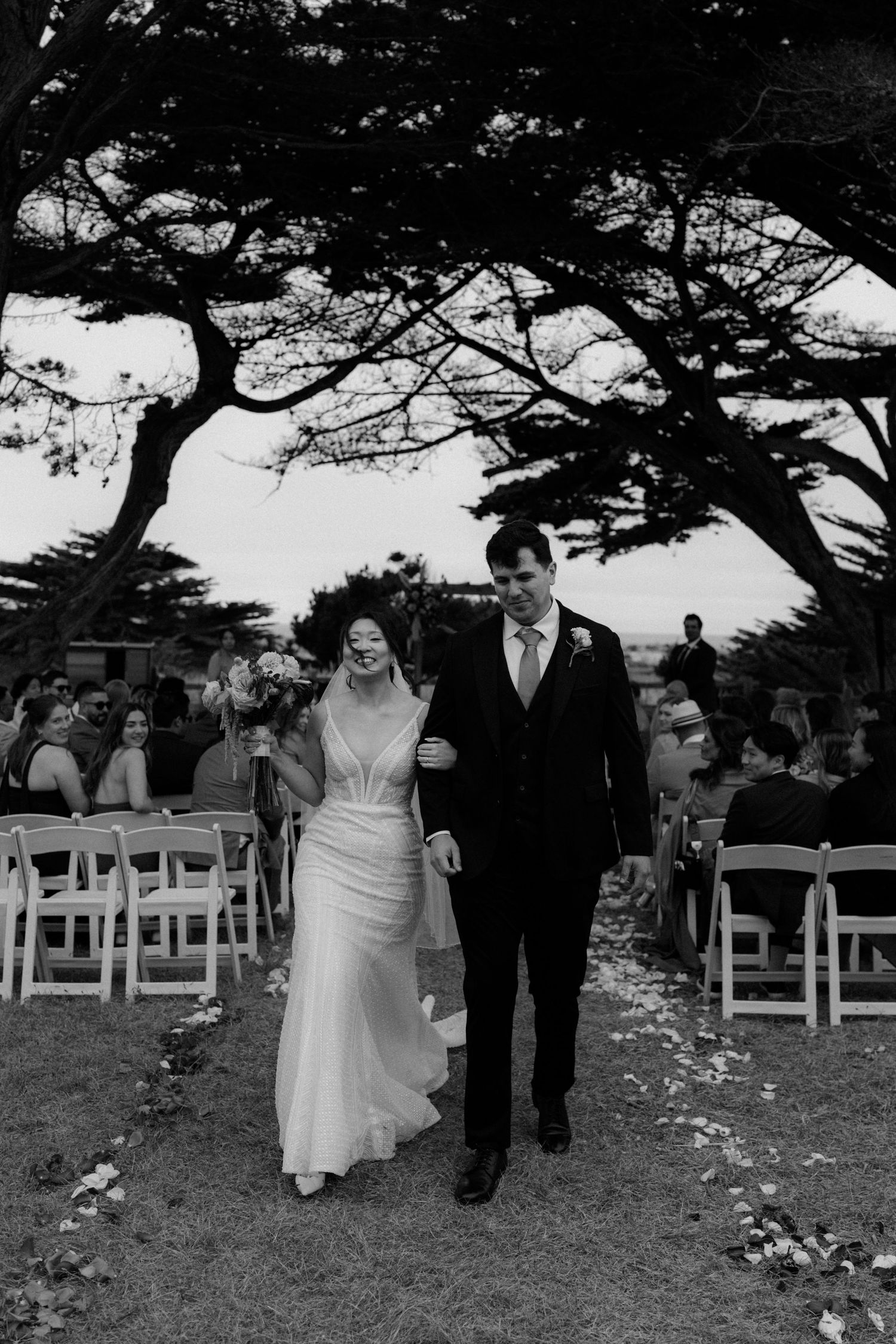Black and white photo of a newlywed couple walking down the aisle after their outdoor wedding ceremony under large trees.