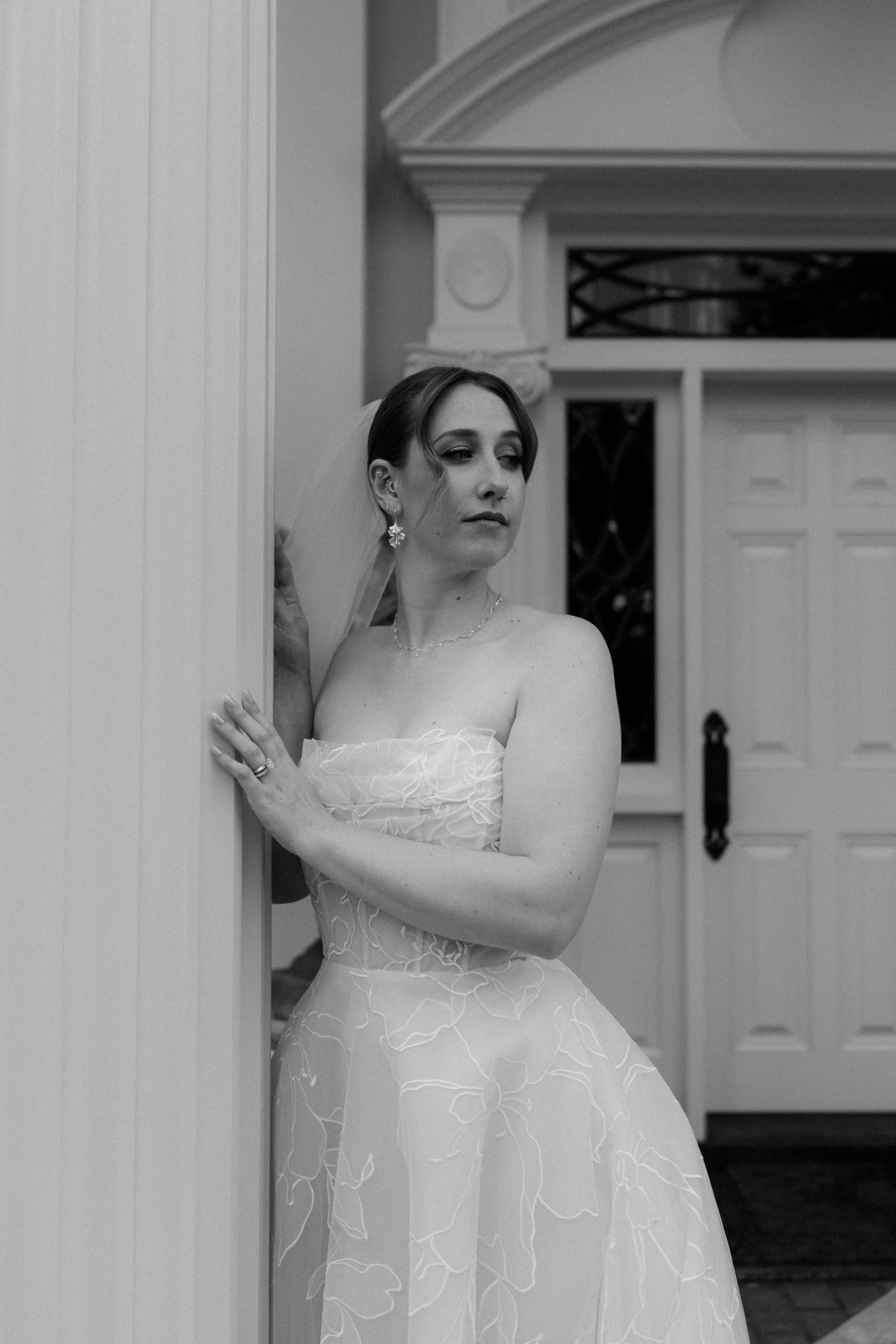 Bride in strapless floral wedding gown leans against white column outside elegant venue, black and white.