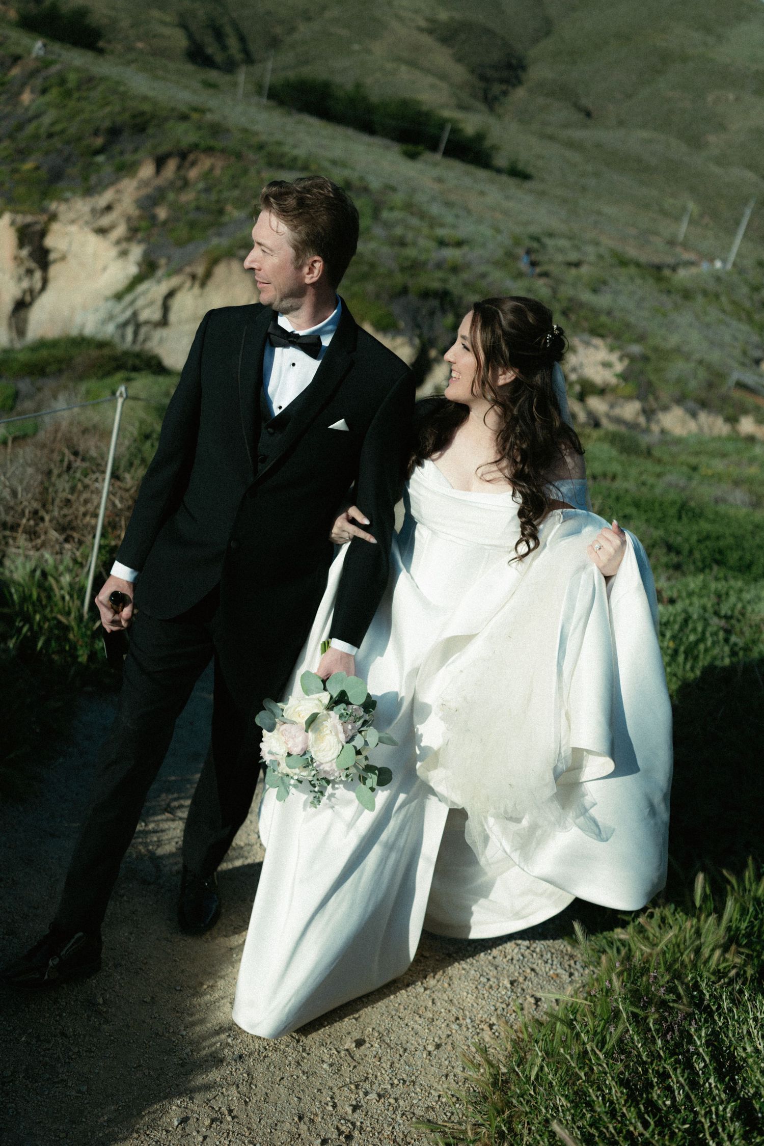 Bride in white off-shoulder gown and groom in black tuxedo pose on a hillside path with green cliffs.