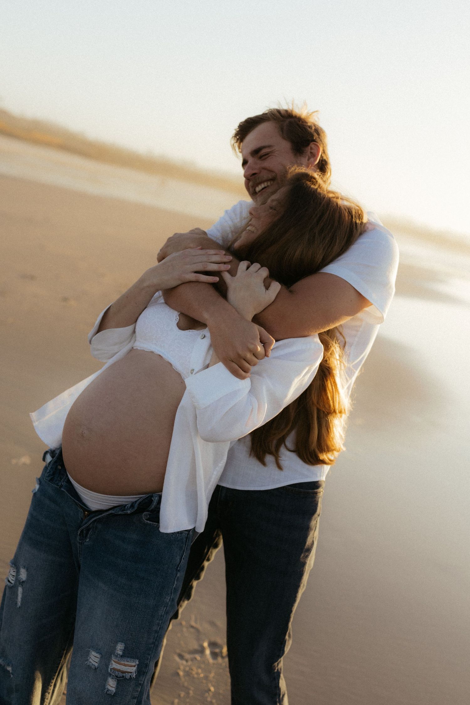 Joyful couple embracing on the beach, with the woman's pregnant belly visible in warm sunset light.