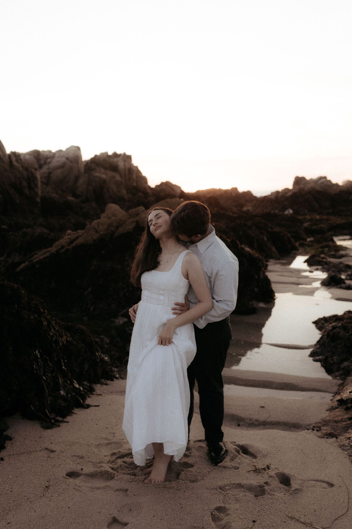 Couple embracing on a sandy beach at sunset, woman in white dress leaning back against man near rocky coastline.