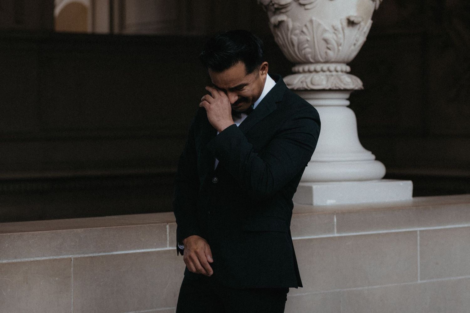 A person in a dark suit stands emotionally near an ornate white pillar.
