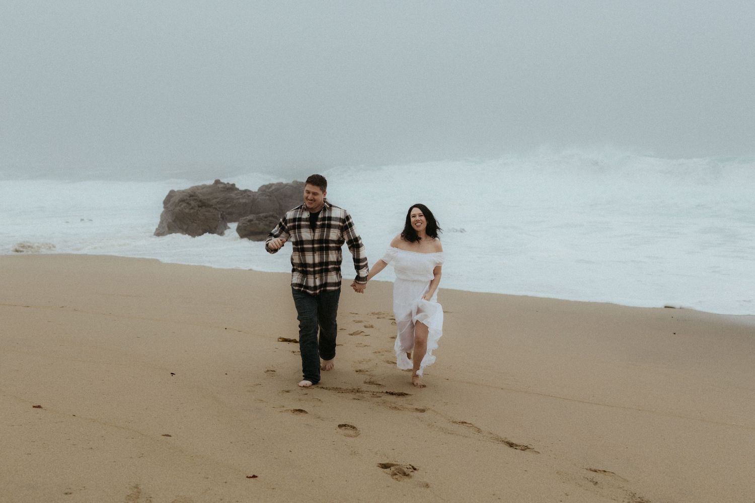 Two people walk along a misty beach shoreline with waves in the background.