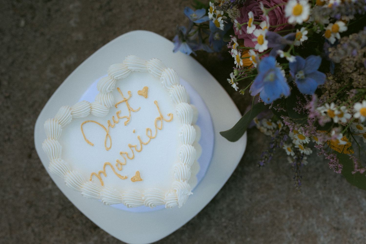Heart-shaped Just Married wedding cake beside wildflower bouquet on stone surface.