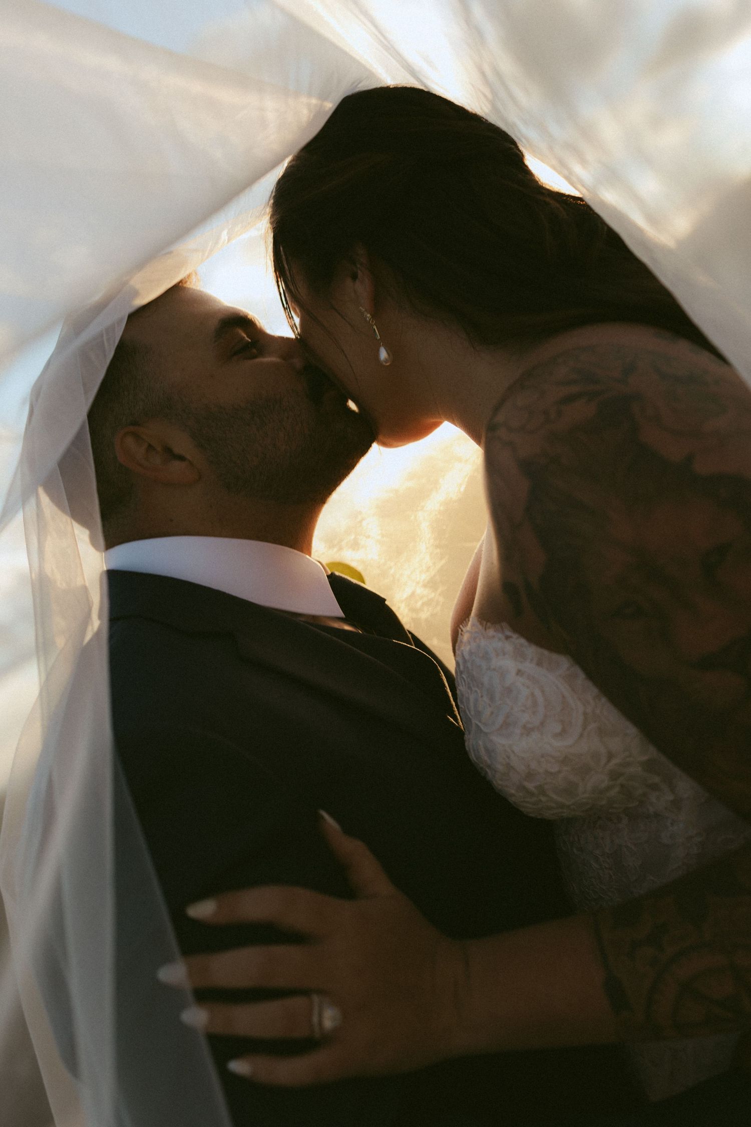 Bride and groom sharing a romantic kiss under a flowing veil with golden backlight on their wedding day.