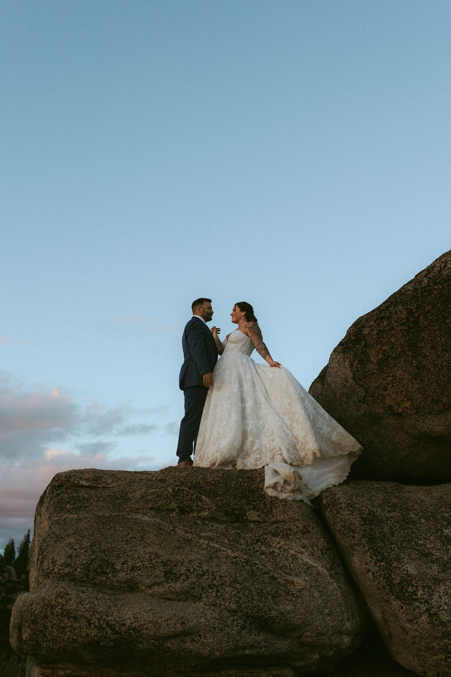 Bride and groom standing on rocky boulder in wedding attire against a blue dusk sky.