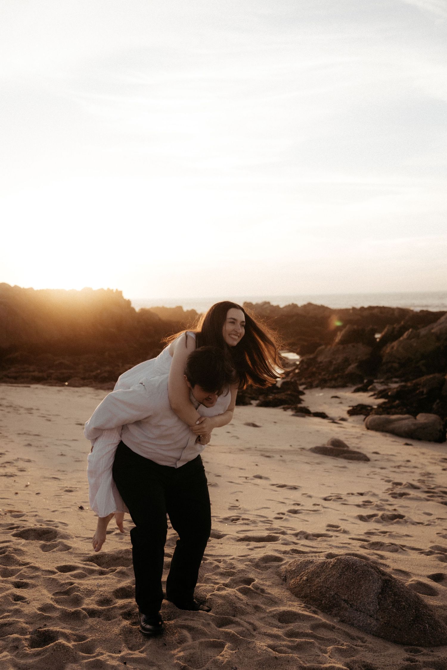 A playful moment captured on a beach at golden hour as a couple shares a piggyback ride among the sand dunes.