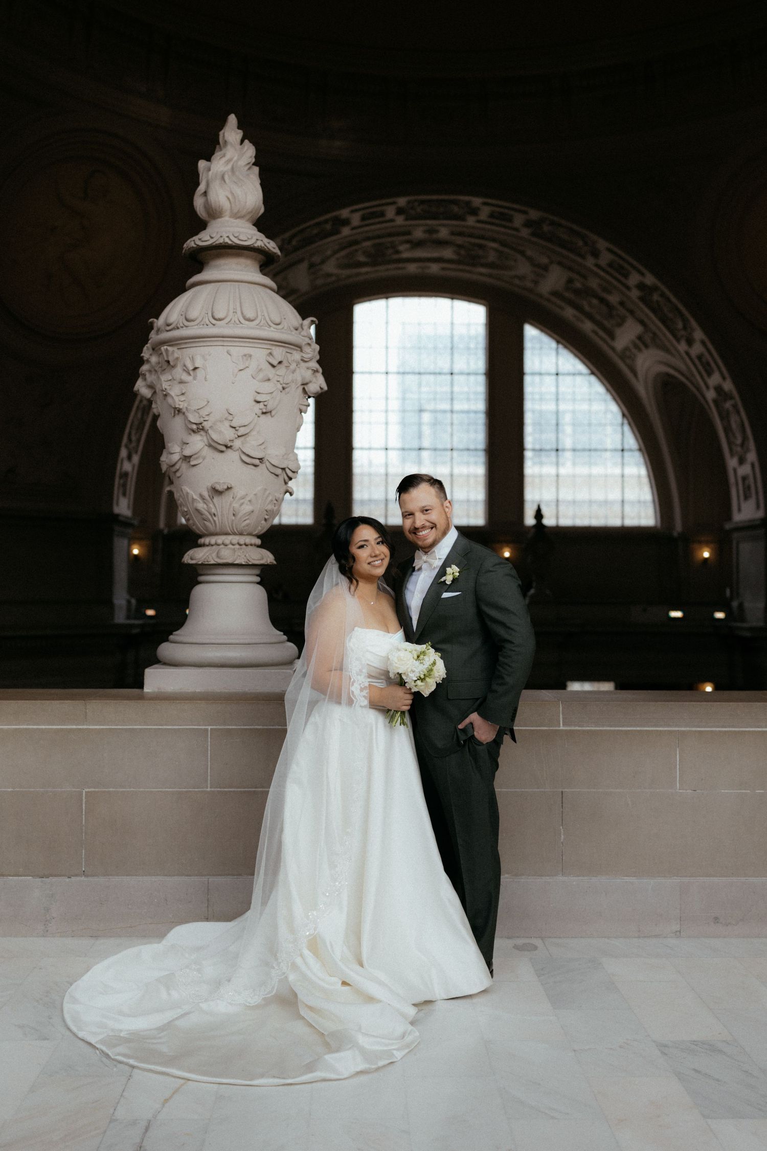 A bride and groom pose for formal wedding portraits in an elegant architectural space with arched windows.