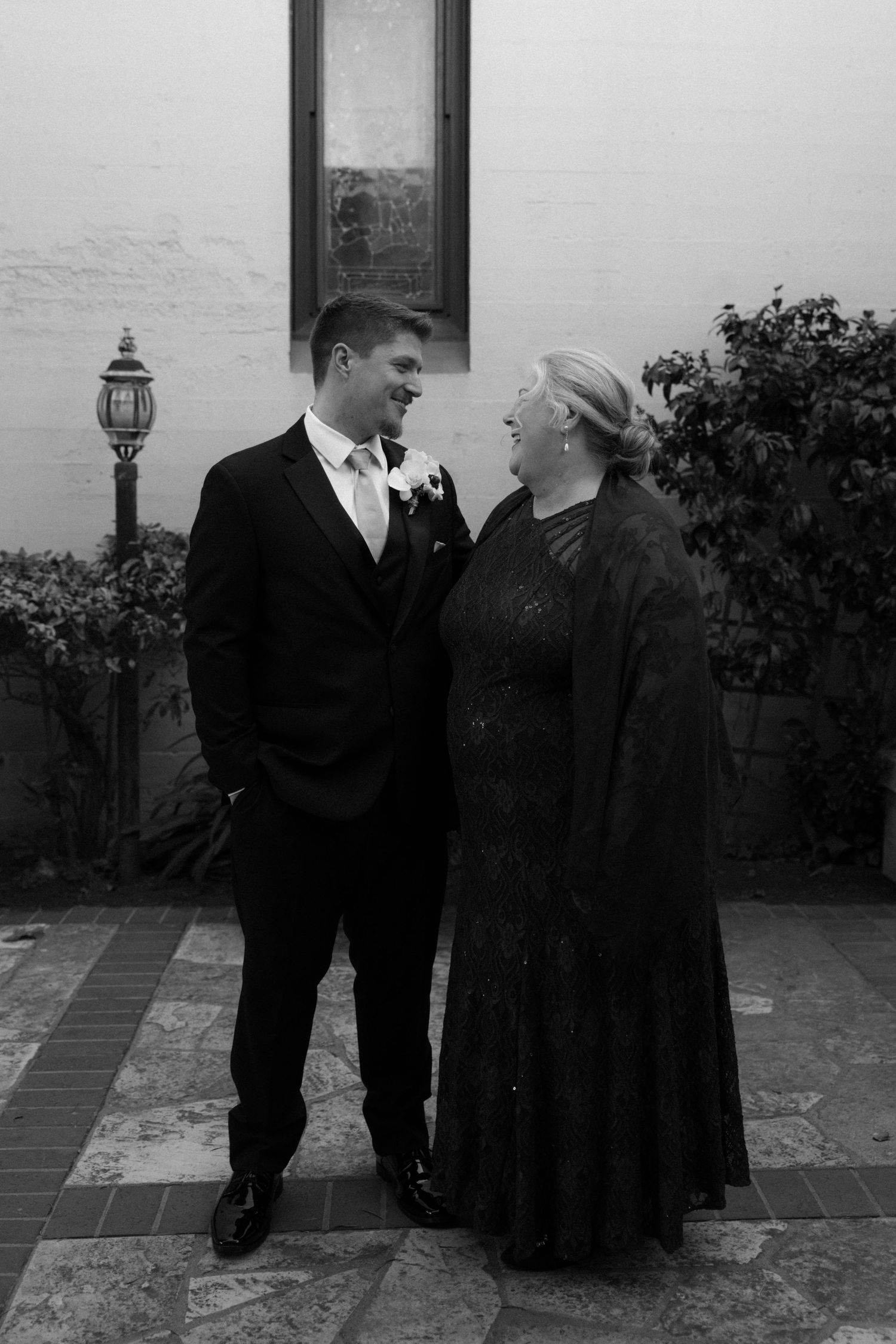 Three people in formal black attire stand together on a stone patio during a wedding event.