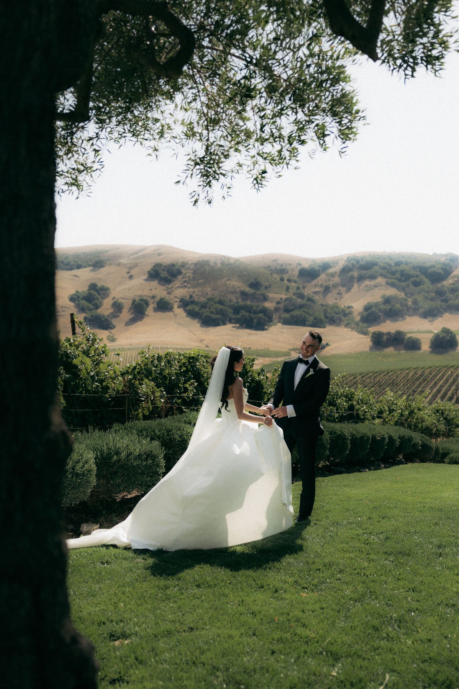 A wedding couple walks across a scenic vineyard lawn with rolling hills in the background.