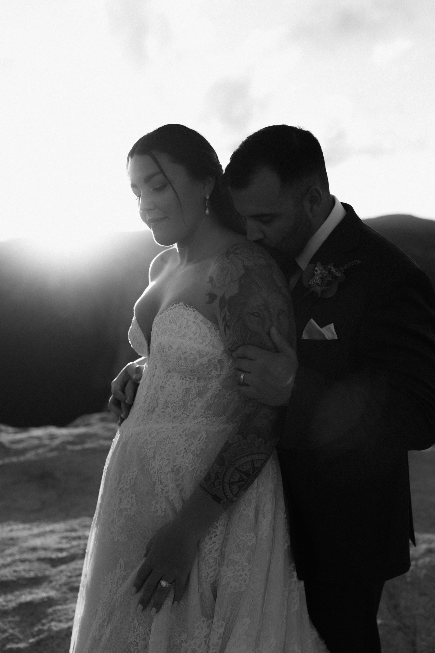 Groom kissing bride on cheek at mountain sunset in romantic black and white wedding portrait.