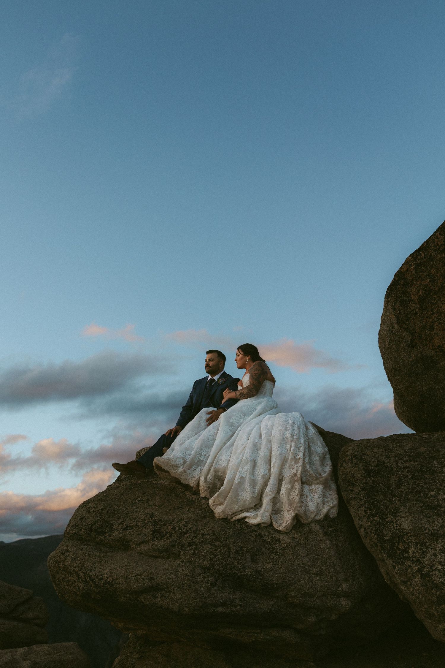 Bride and groom sitting on rocky summit at twilight with dramatic blue sky backdrop during outdoor elopement.