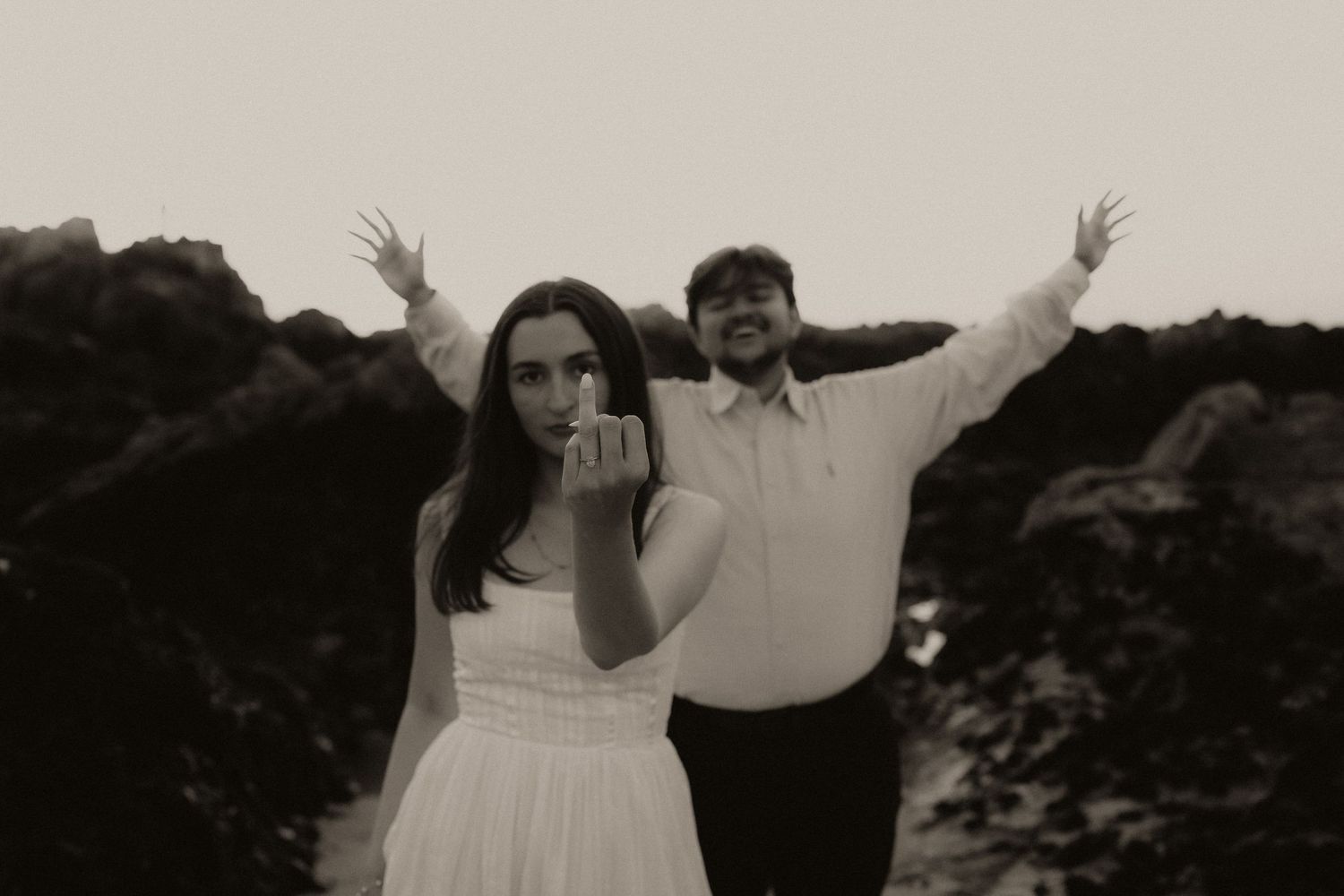Black and white photo of couple posing on rocks, woman showing ring, man with arms raised.
