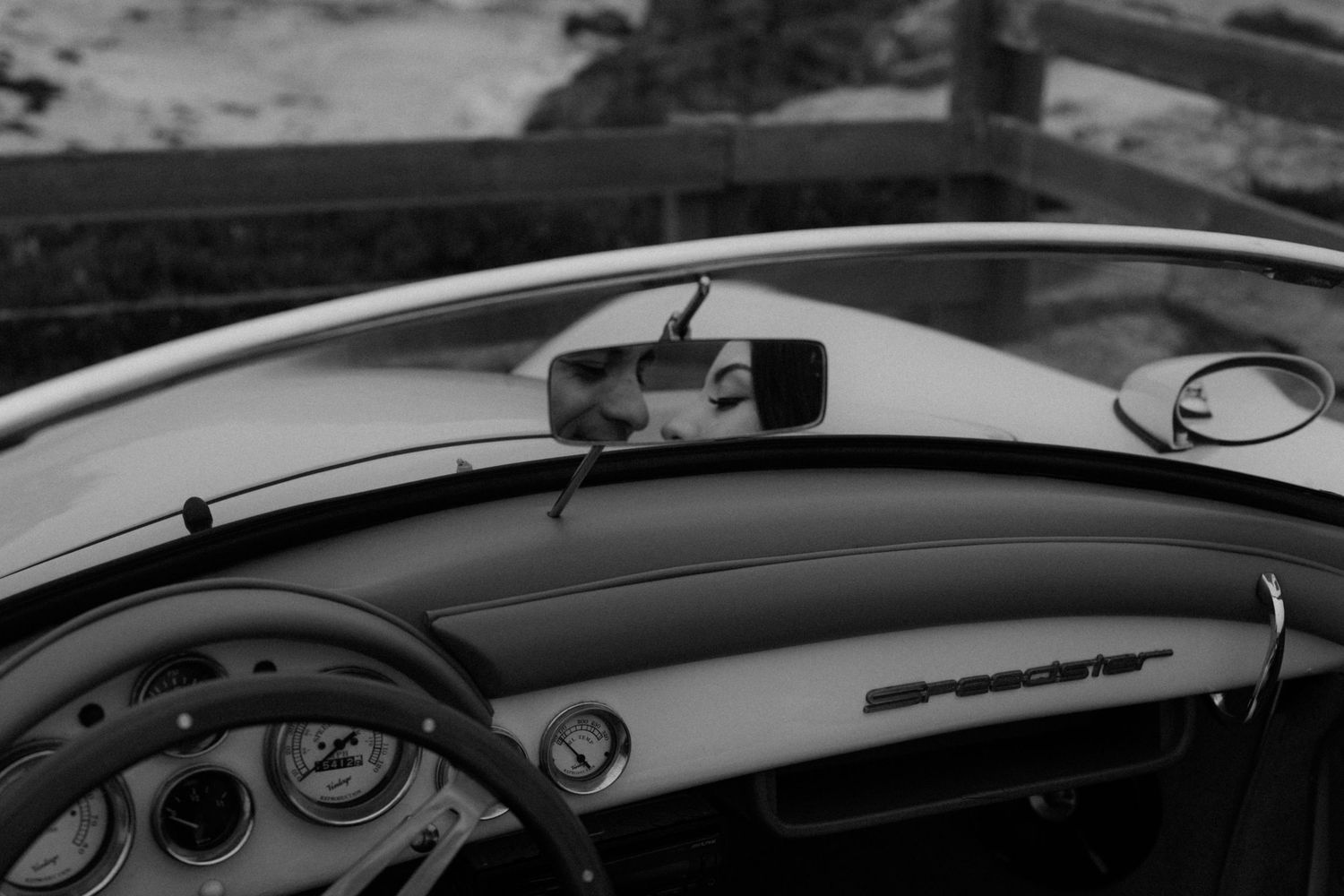 Black and white photo of a vintage car interior dashboard with rearview mirror reflecting a person.