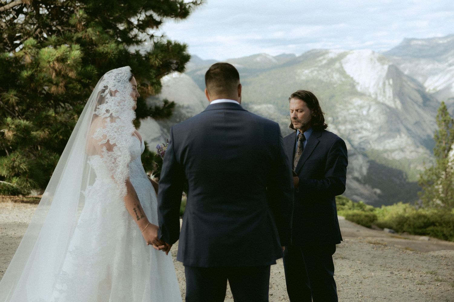 Bride and groom hold hands during outdoor wedding ceremony with stunning Yosemite mountain backdrop.