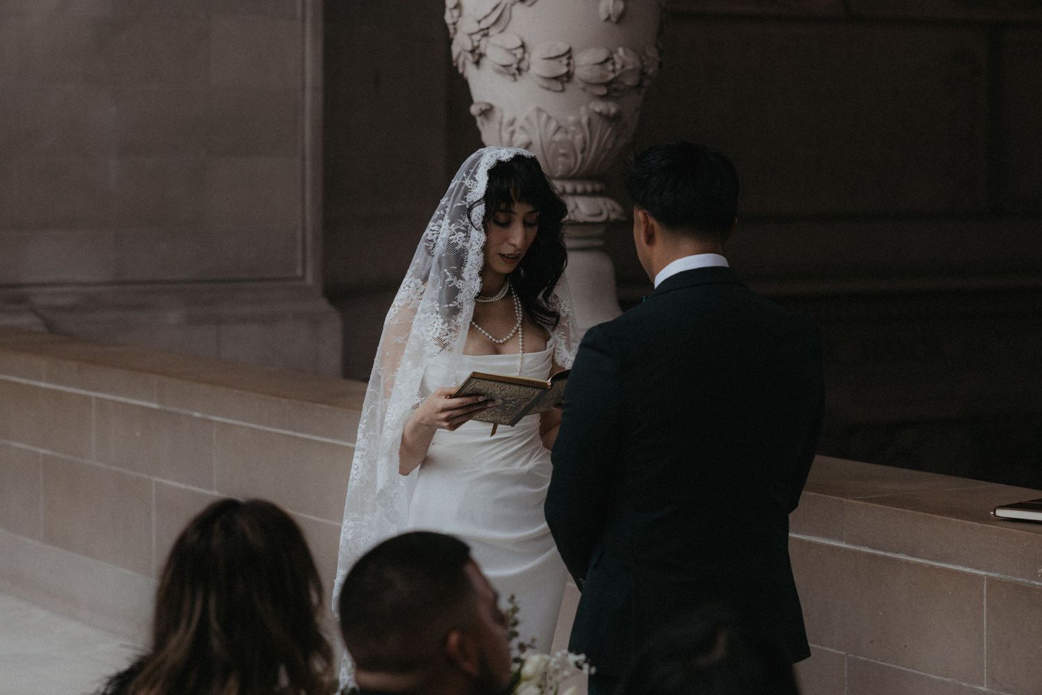 Bride in white gown and lace veil reads wedding vows during an elegant indoor ceremony.