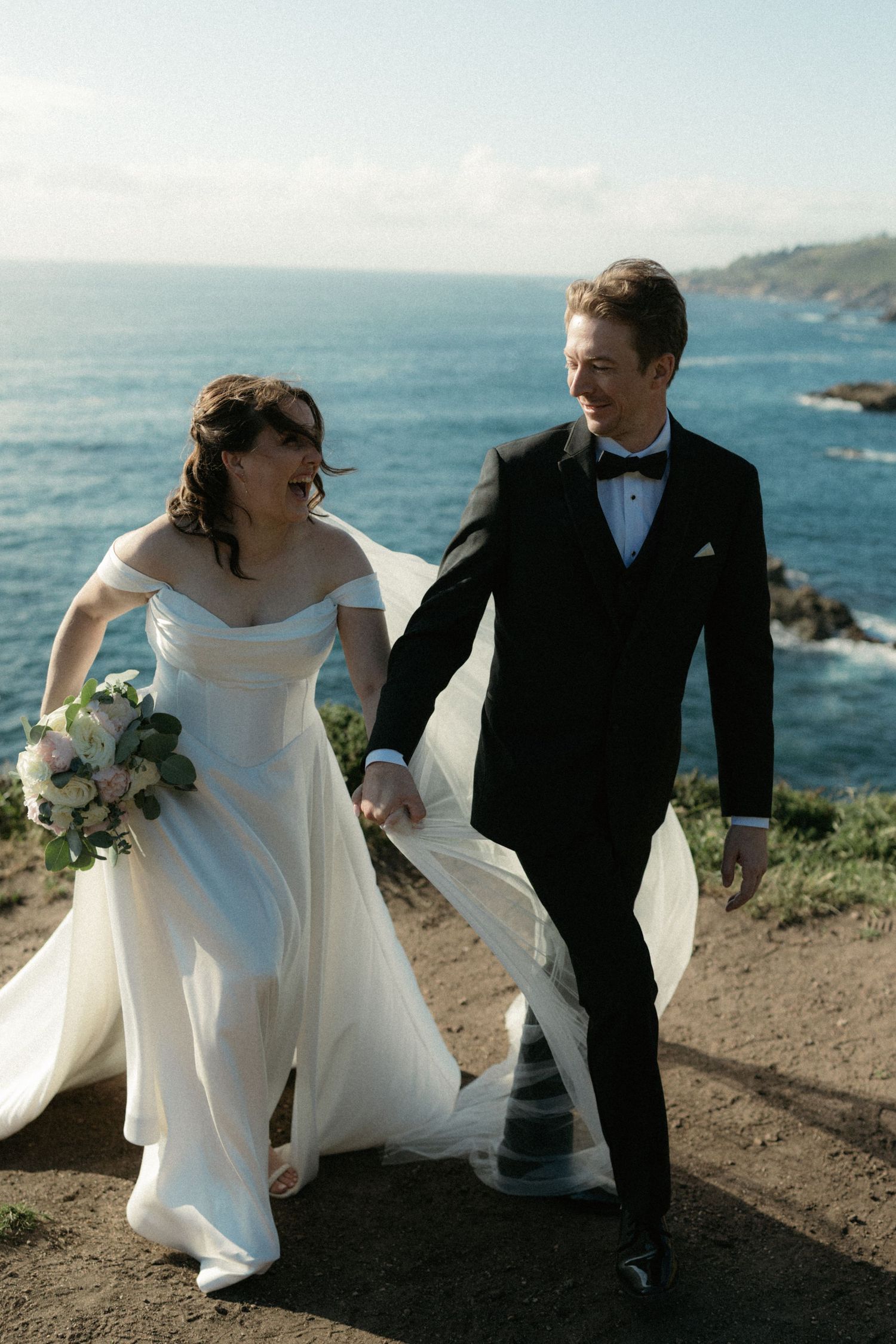 Smiling bride and groom walk hand-in-hand on a coastal cliff with ocean views on their wedding day.