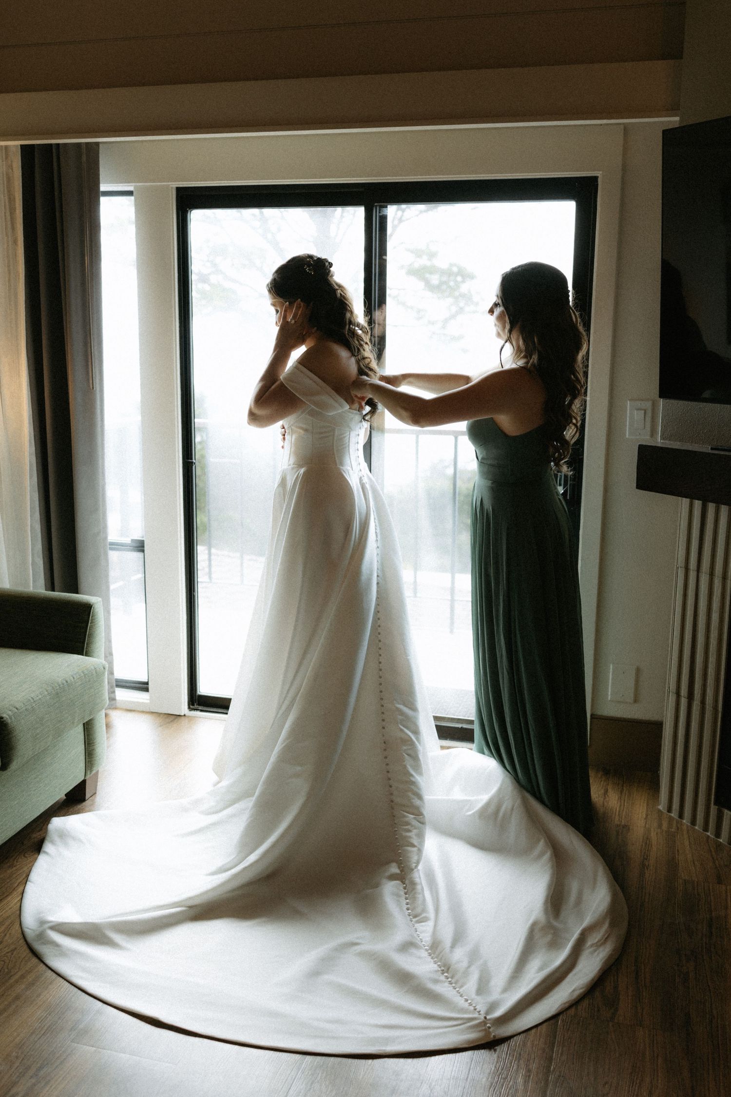 Bridesmaid in green dress helping bride with her white wedding gown in a hotel room by large windows.