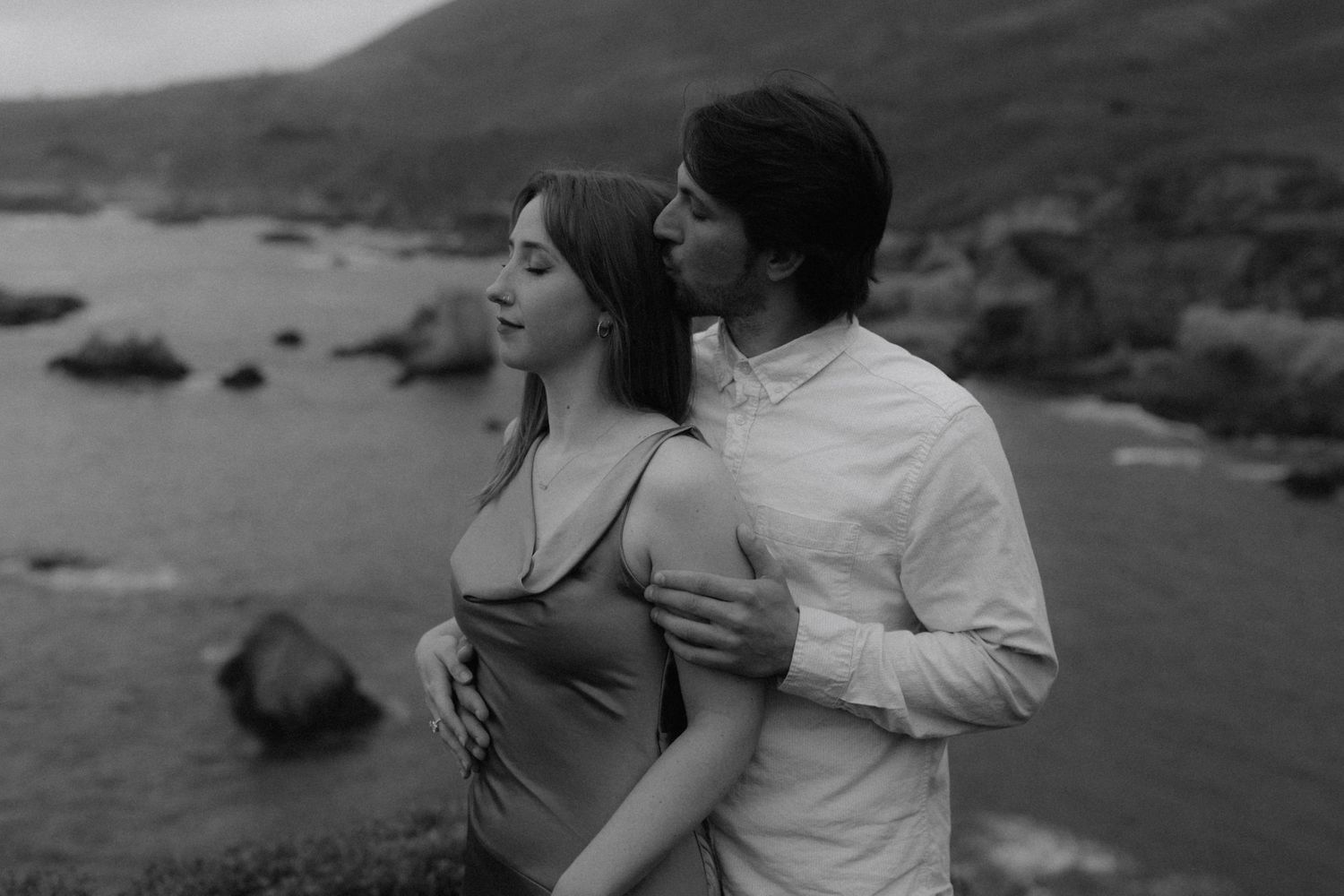 Black and white photo of a couple embracing near a rocky coastal shoreline, the man holding the woman from behind.