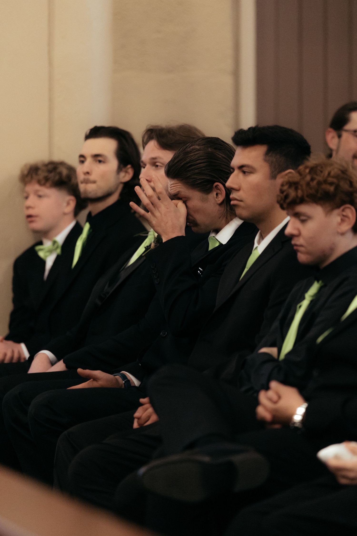 Groomsmen in black suits with light green ties seated in a row at a church ceremony.