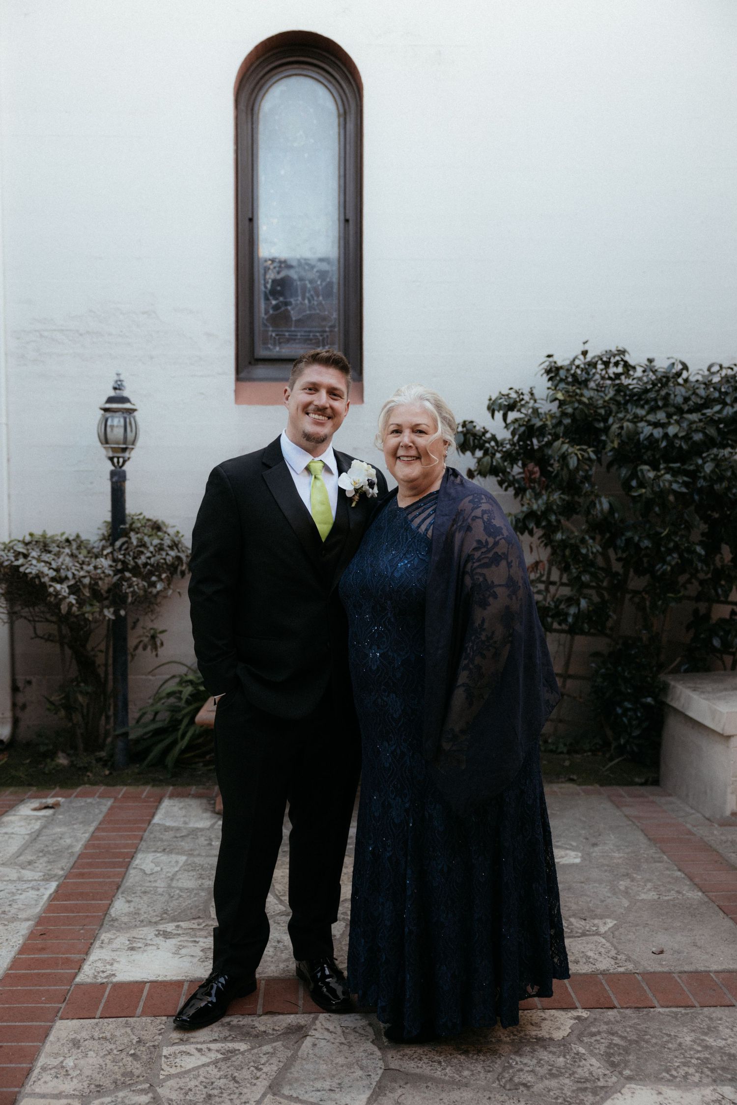 Two people in formal attire stand together on a tiled patio with arched windows and greenery in the background.