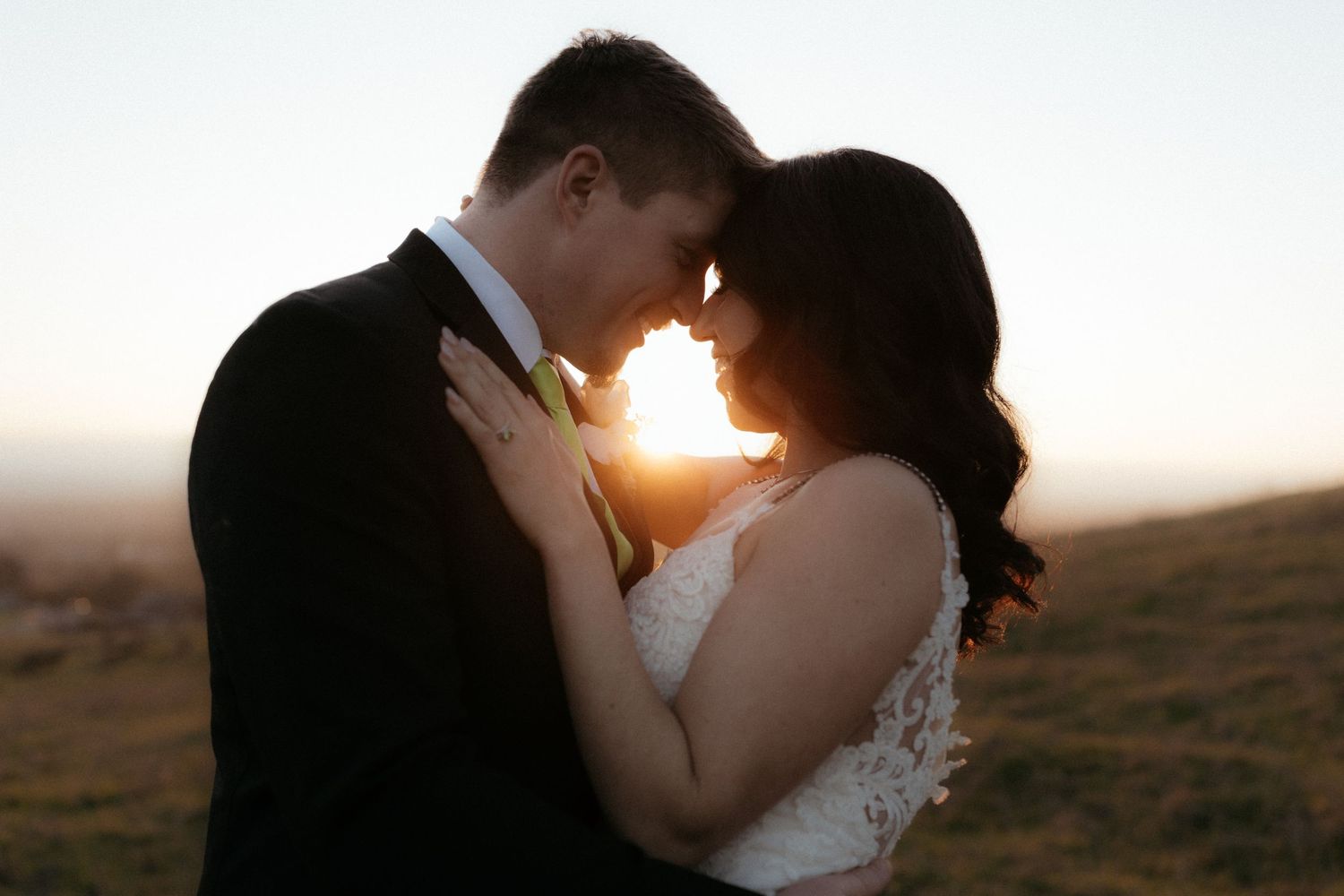 A couple shares a romantic kiss at sunset in a scenic outdoor location with golden light streaming through the background.